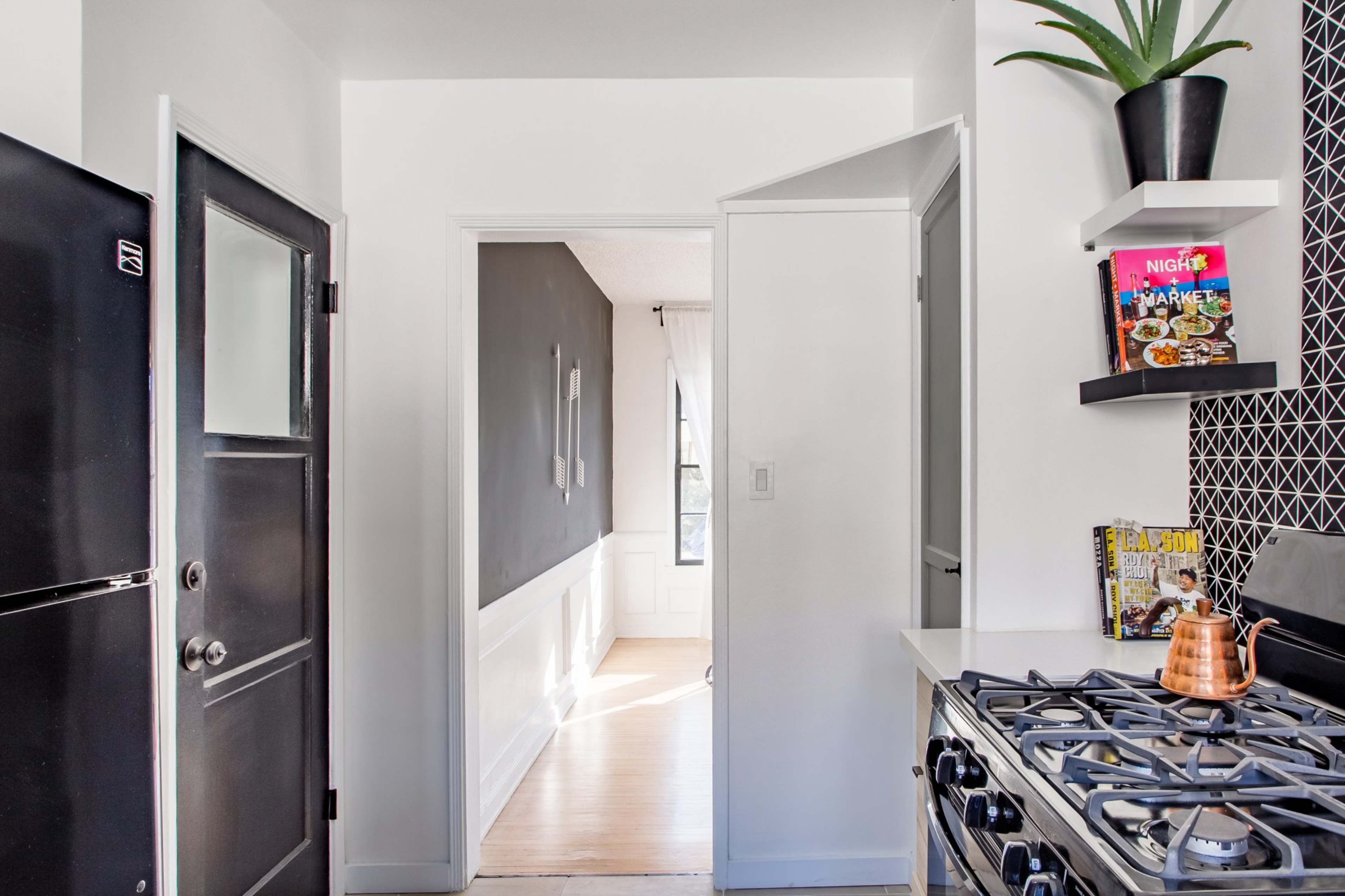 A modern kitchen with a view through a doorway into a bright hallway, featuring a black refrigerator and a gas stove.