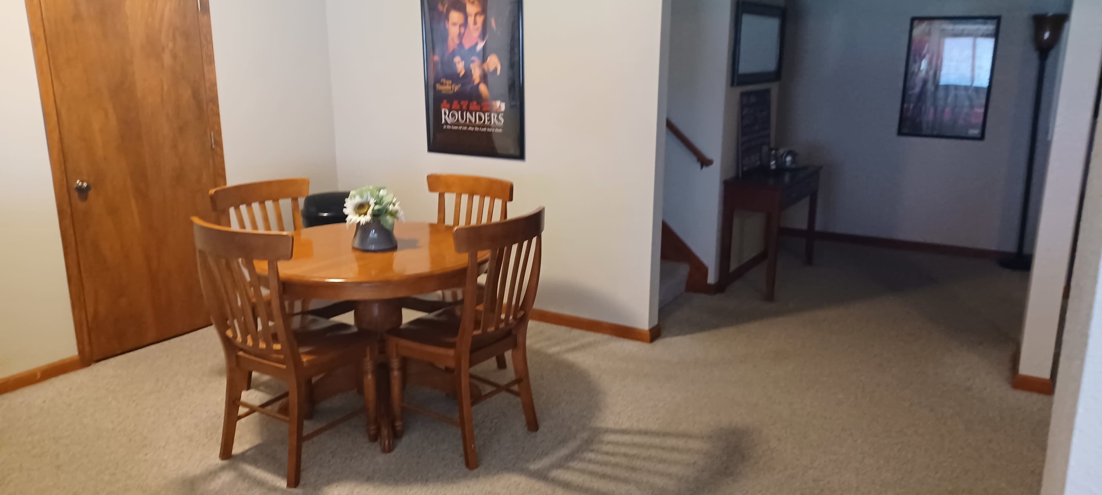 A wooden dining table with four chairs sits near a wall adorned with a movie poster, adjacent to a hallway leading to another room.