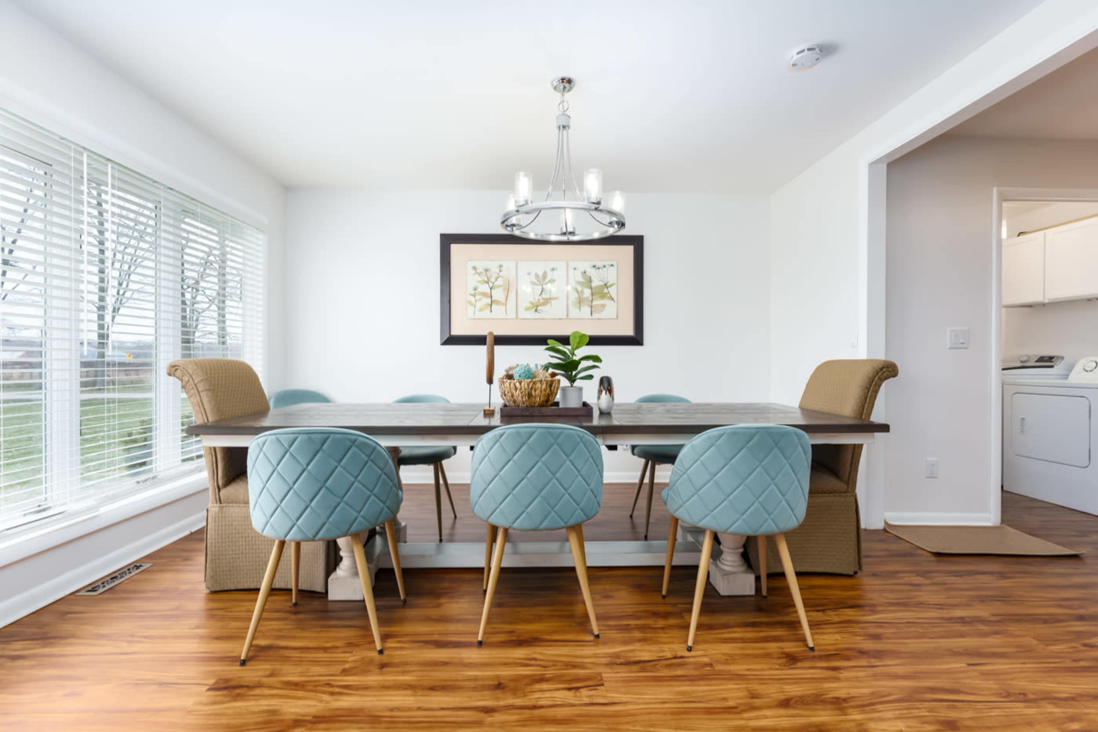 A modern dining room features a long wooden table surrounded by six quilted chairs, with a chandelier above and large windows allowing natural light.