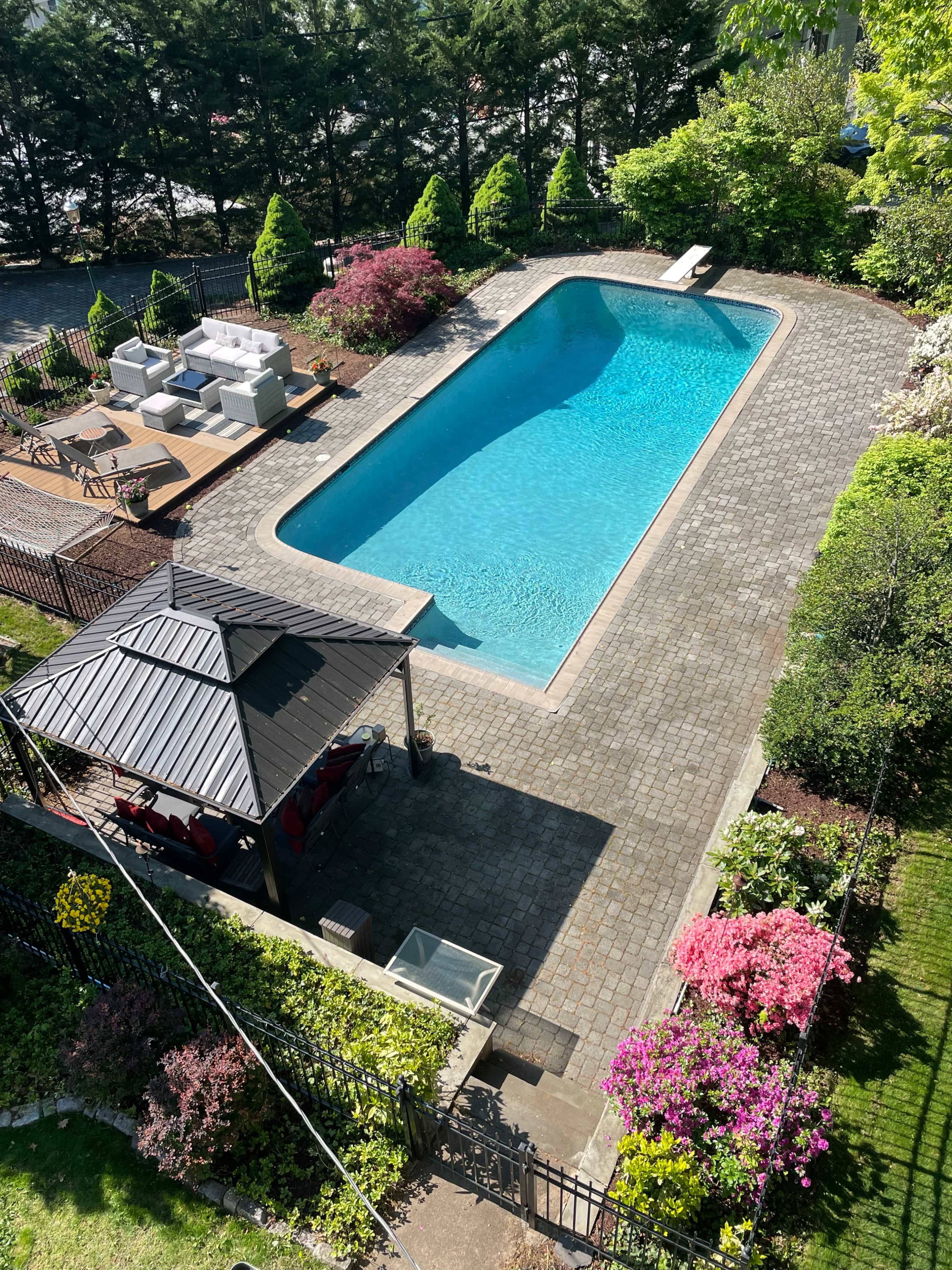 The image shows a rectangular swimming pool surrounded by blooming flowers and greenery, with a gazebo and outdoor seating area nearby.