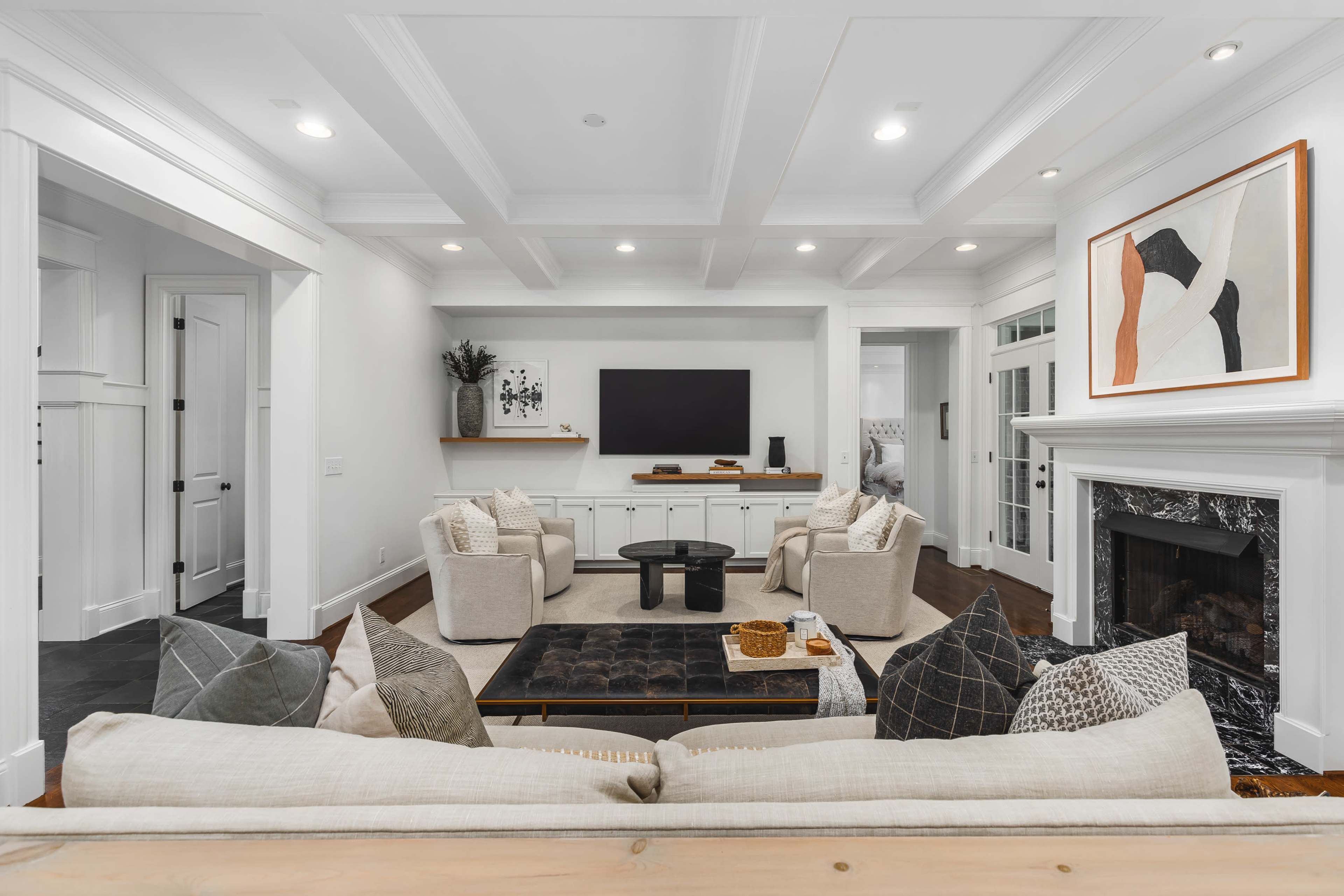 A modern living room features a central coffee table surrounded by plush armchairs and a fireplace, with a television mounted on the wall and artwork displayed nearby.