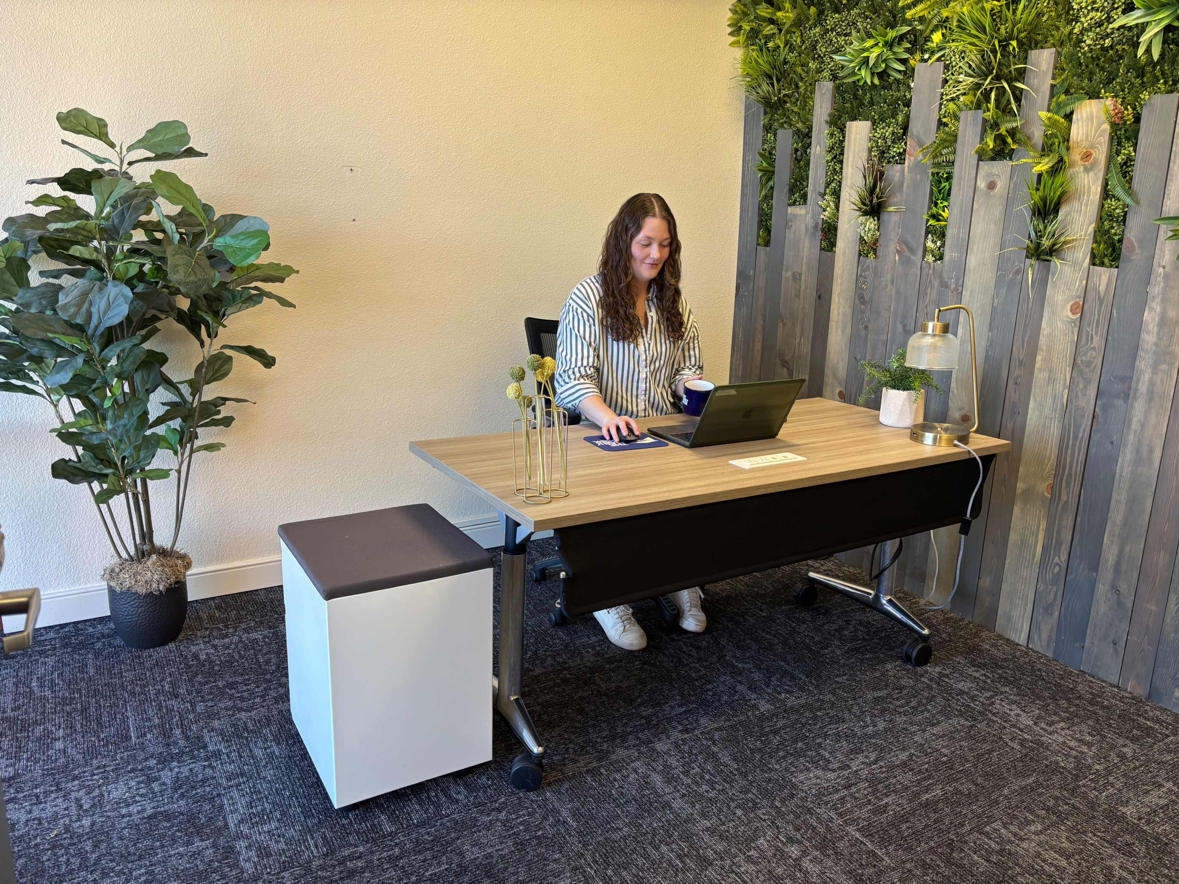 A person sits at a modern desk working on a laptop in an office space with greenery and wooden accents in the background.