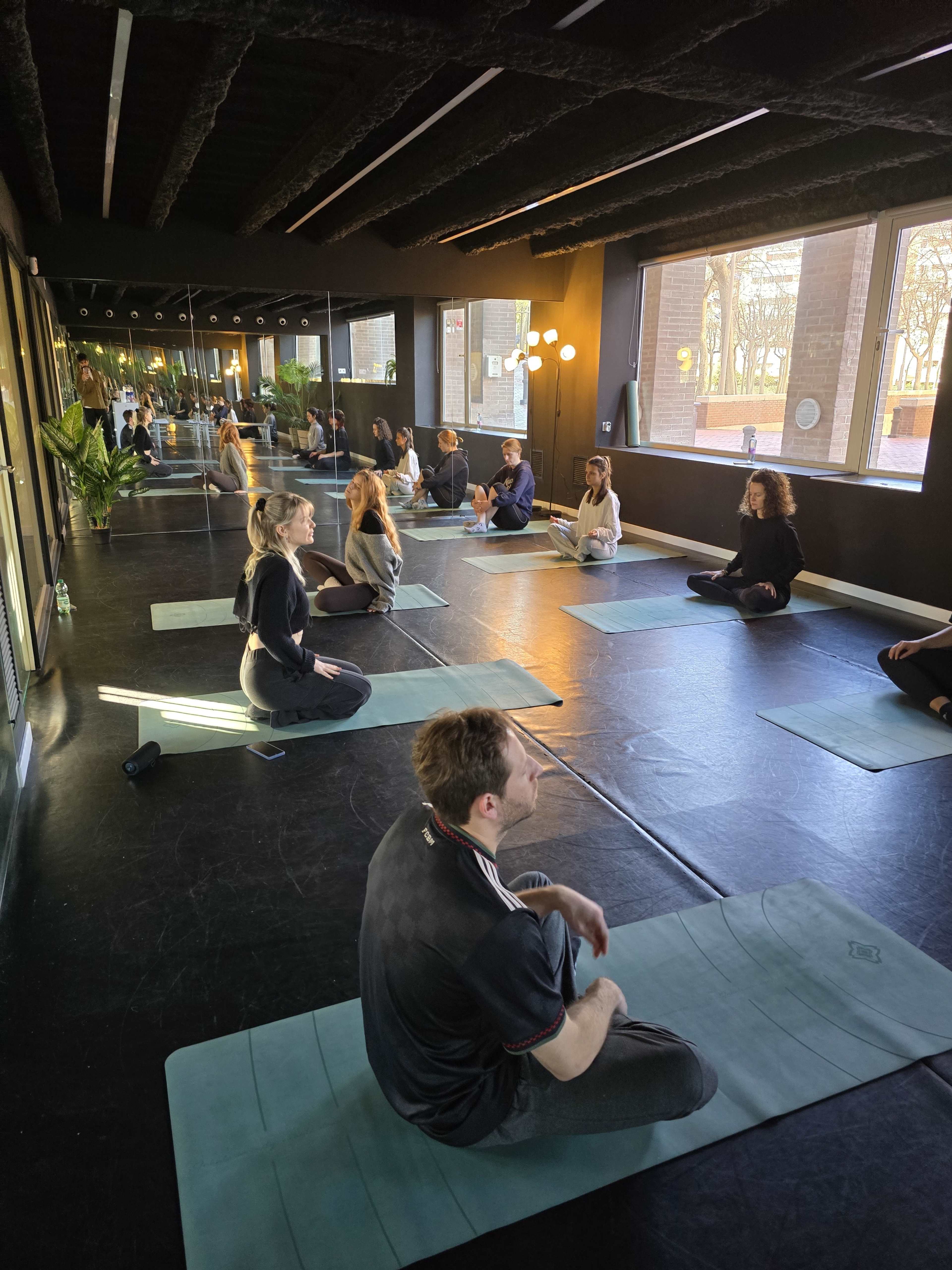 People are seated on yoga mats in a spacious studio with large windows and mirrored walls.