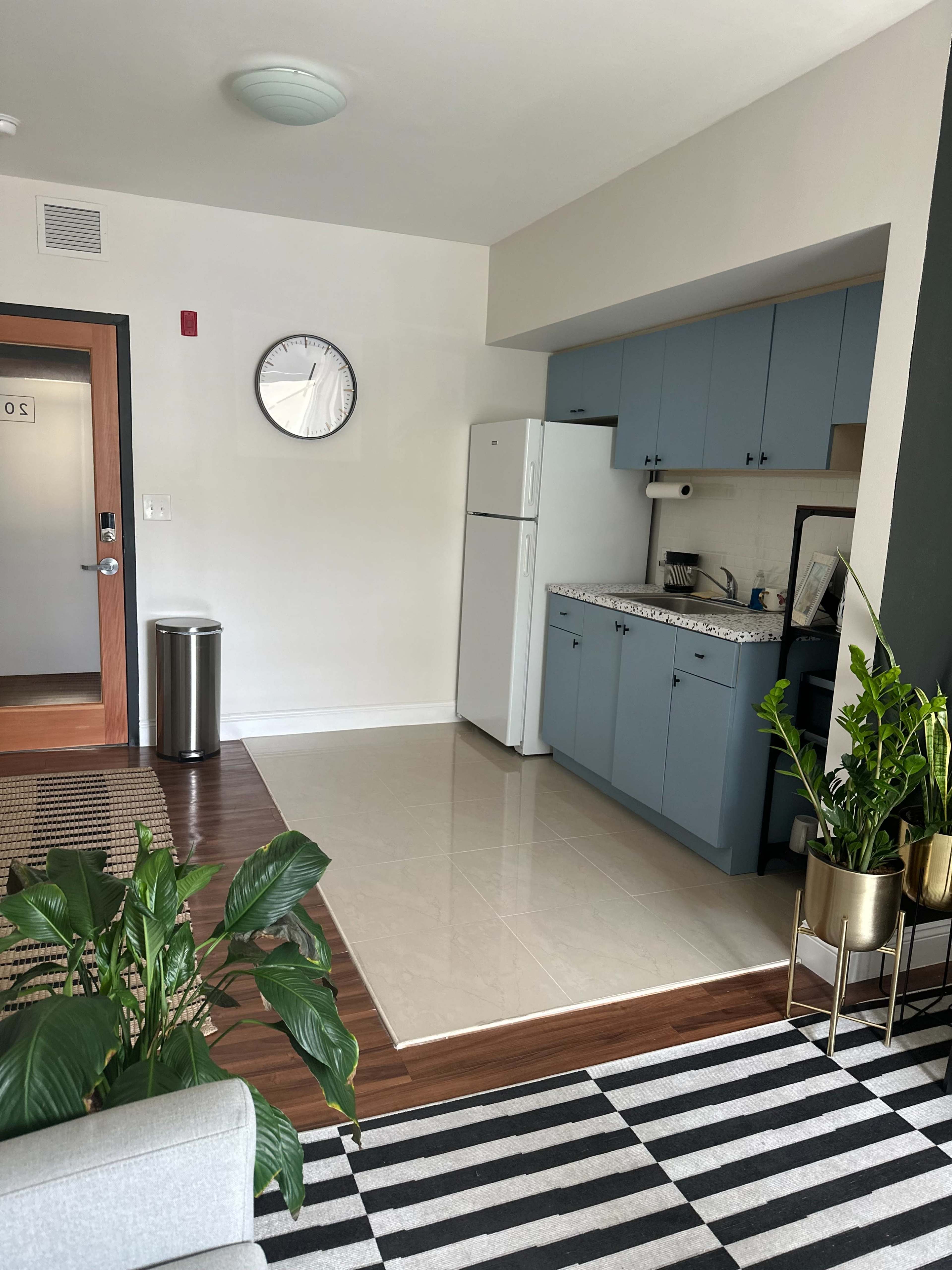 A modern kitchen area with blue cabinets, a white refrigerator, and a clock on the wall, adjacent to a living space featuring a striped rug and potted plants.