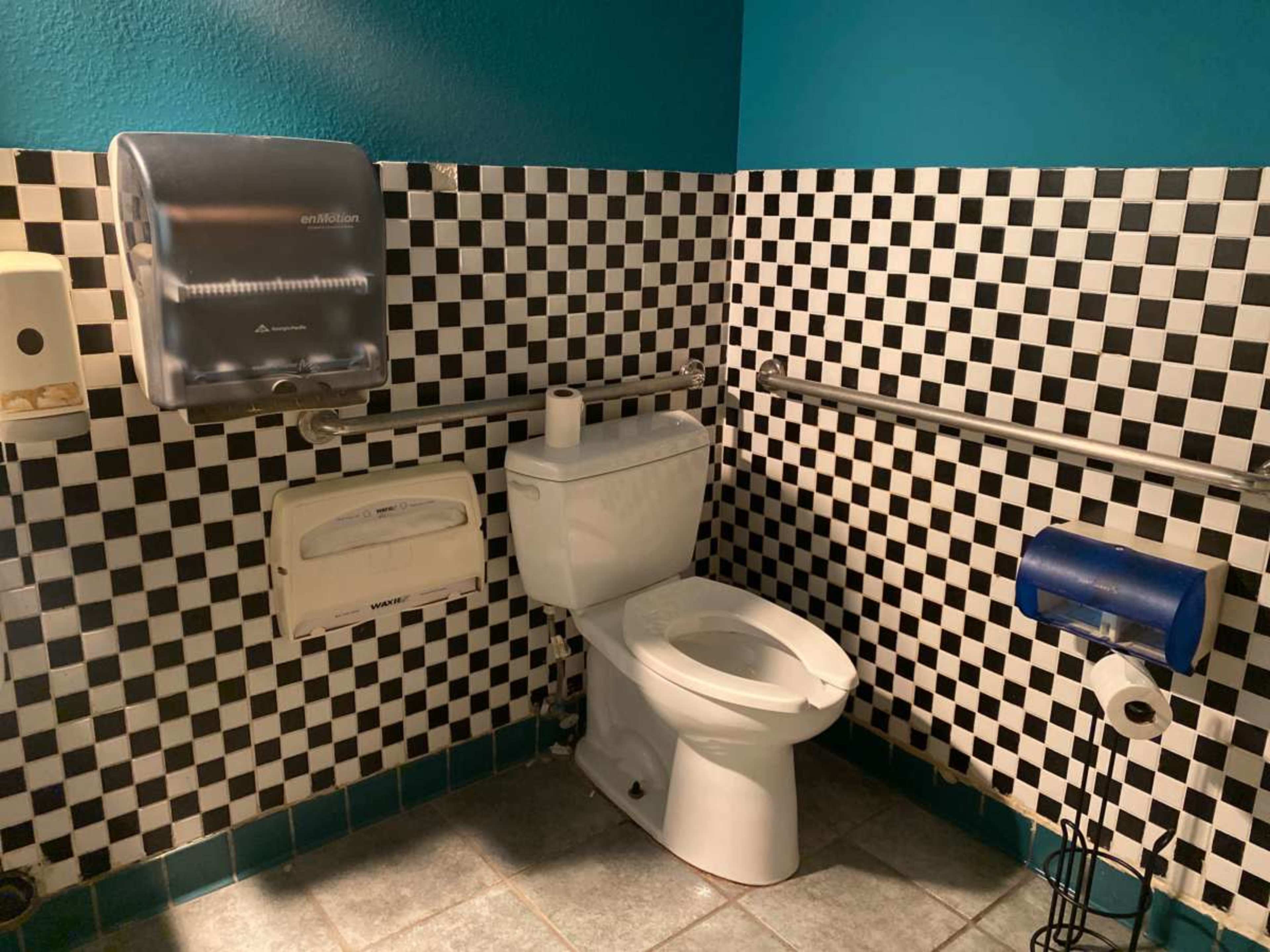 The image shows a bathroom stall featuring a white toilet against a backdrop of black and white checkered tiles, with various dispensers and a toilet paper holder nearby.