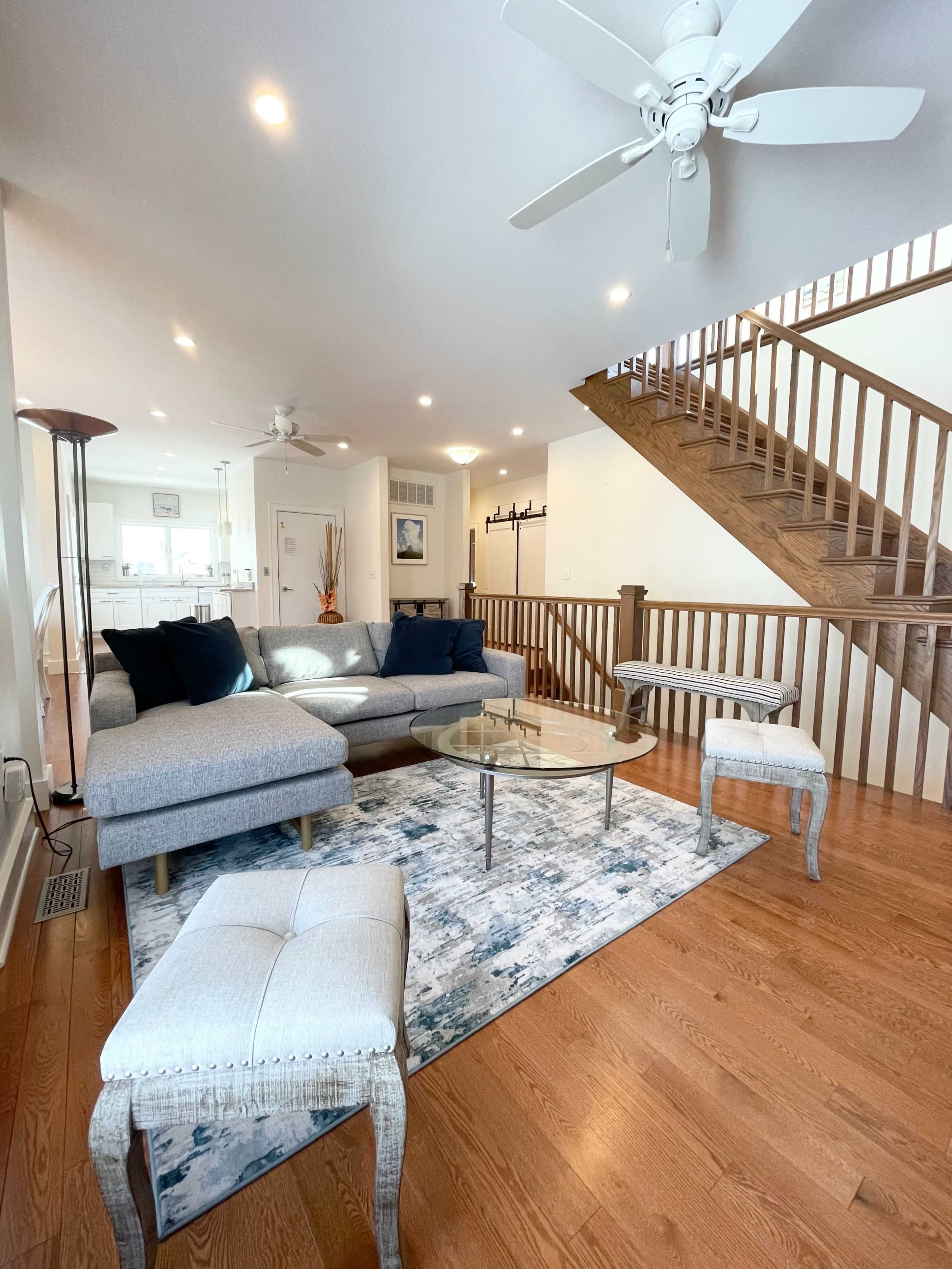 A modern living room with a gray sectional sofa, a glass coffee table, and a staircase in the background.