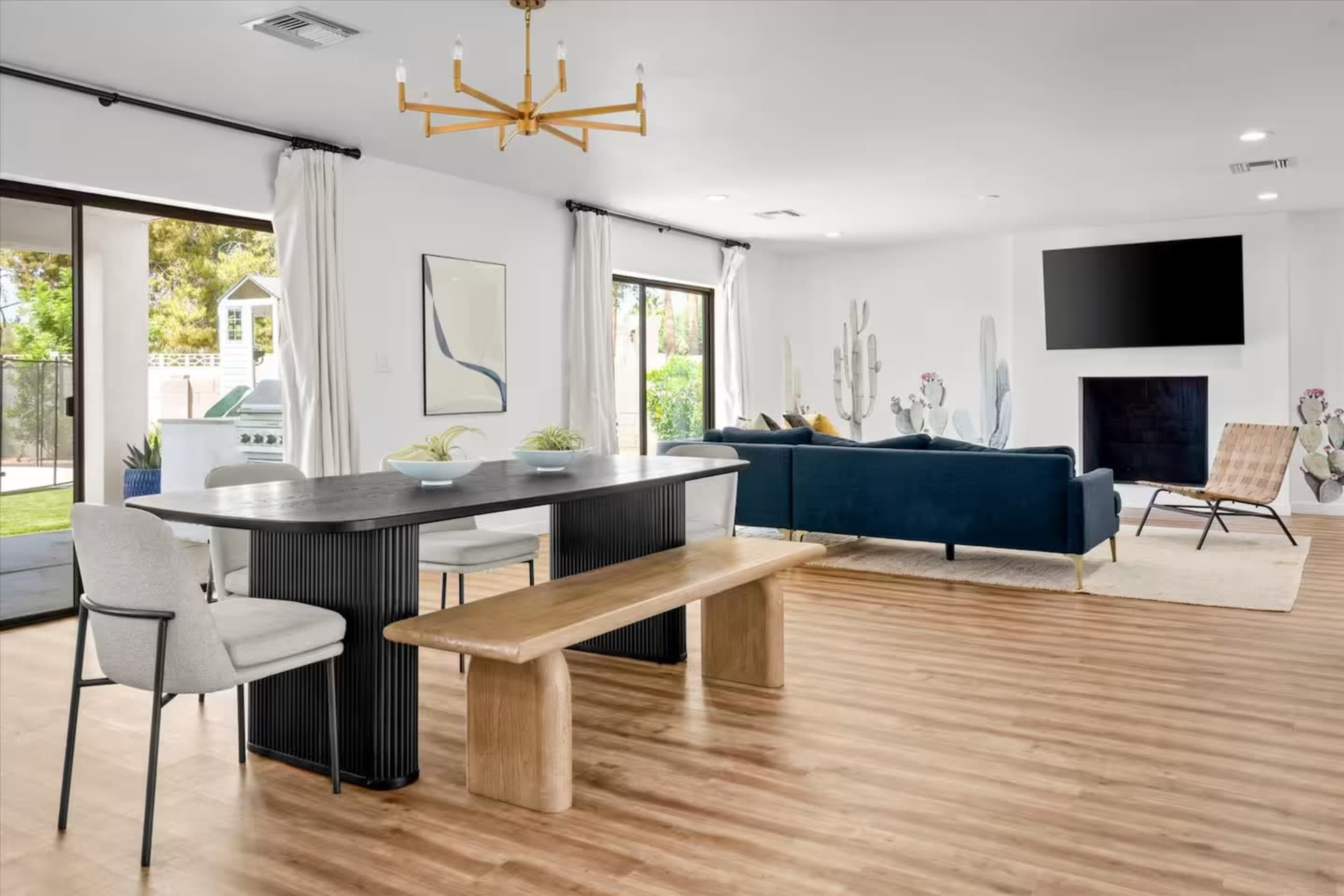 The image shows a modern living space featuring a dining table with two chairs, a wooden bench, a blue sofa, a large television mounted on the wall, and large windows overlooking a garden.