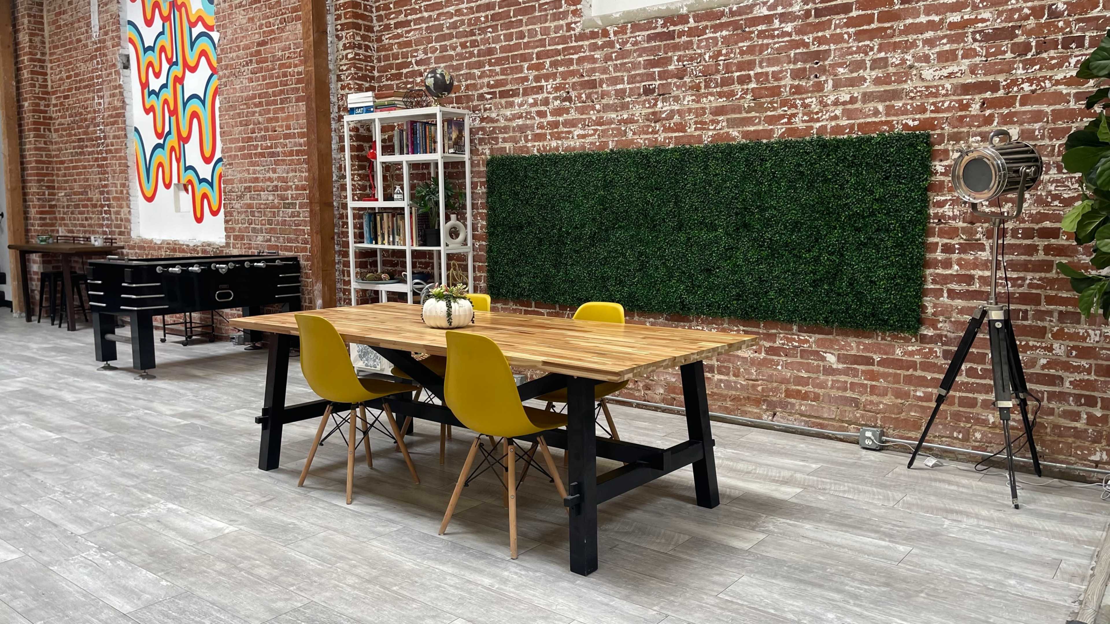 A wooden table with yellow chairs is set in a spacious room with exposed brick walls and a green wall panel.
