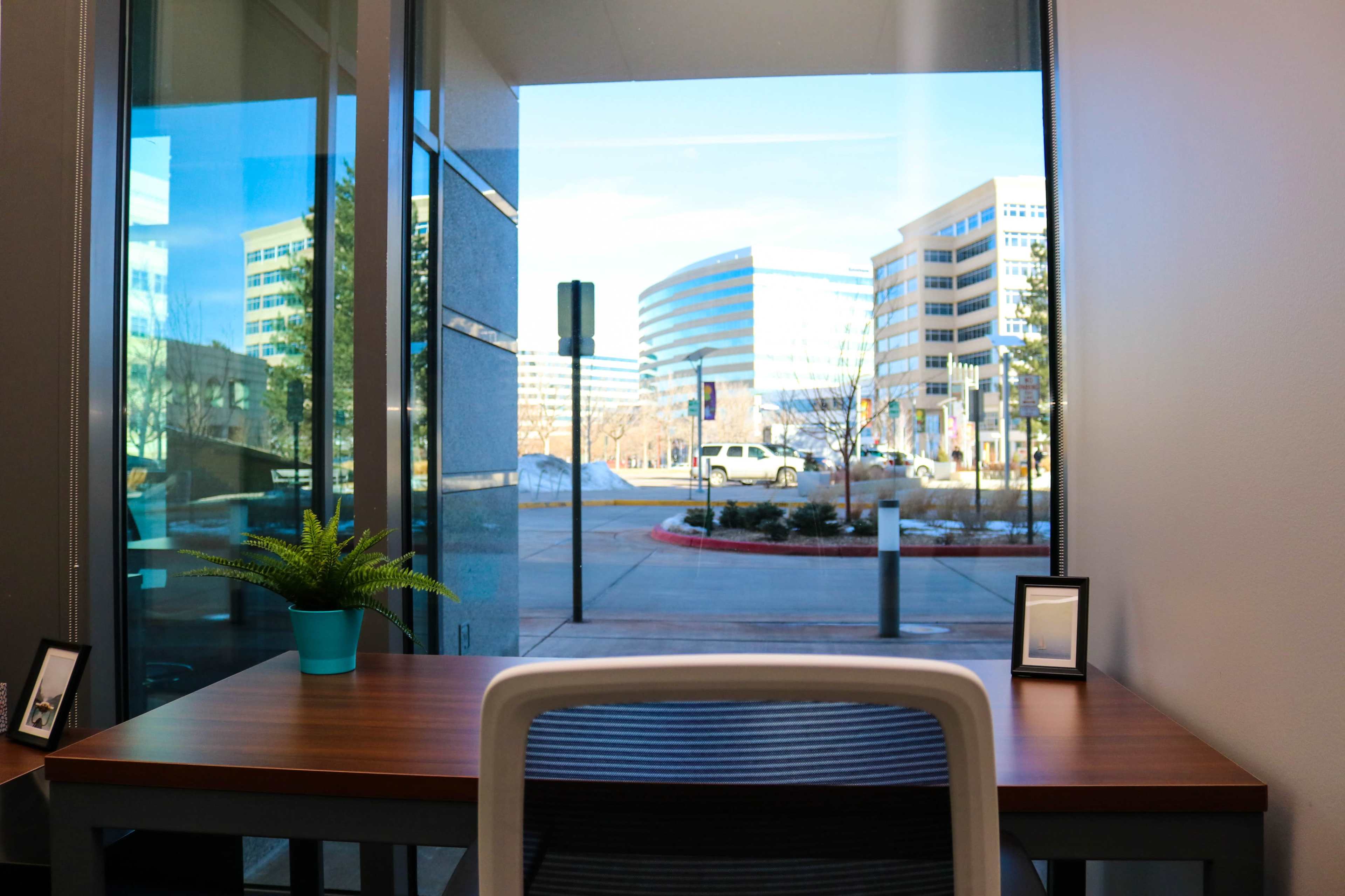 A modern workspace features a wooden table with a potted plant and two picture frames, overlooking a city street through large glass windows.
