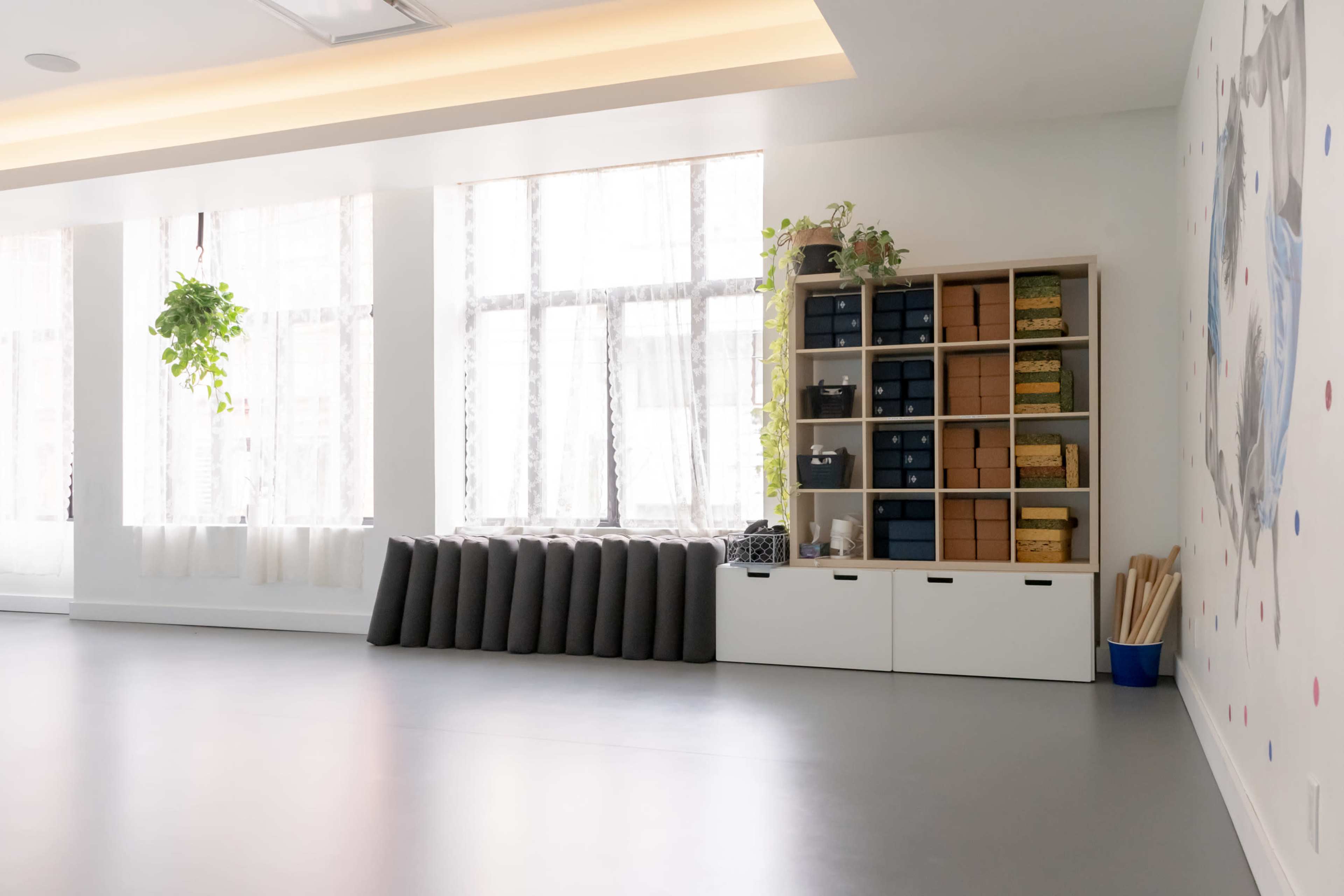 A bright, minimalist room with large windows, a shelf filled with storage boxes, and neatly arranged yoga mats.