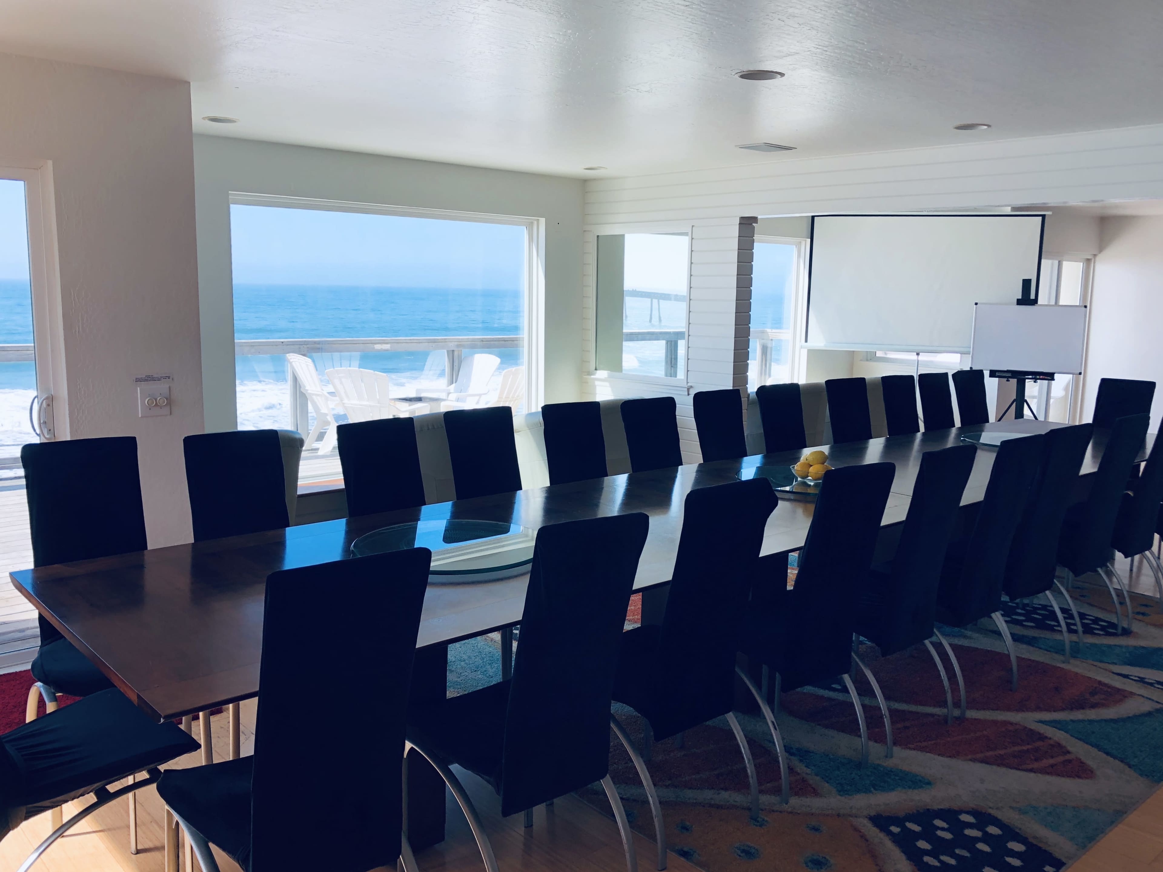 A long conference table with black chairs is set up in a brightly lit room overlooking the ocean, featuring large windows and a projector screen.