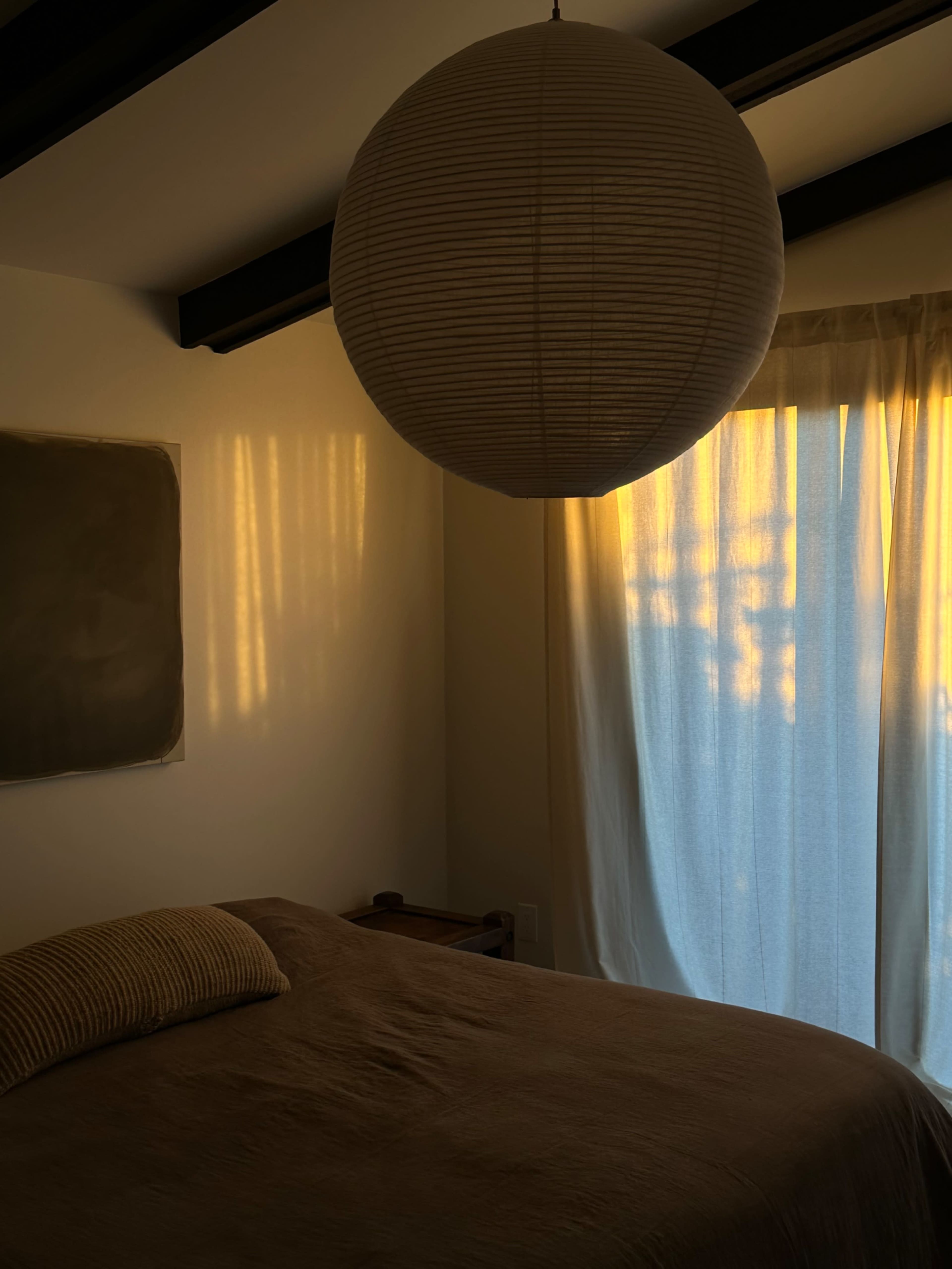 A bedroom features a bed with a beige throw pillow, a lantern hanging from the ceiling, and sunlight casting shadows on the wall through sheer curtains.