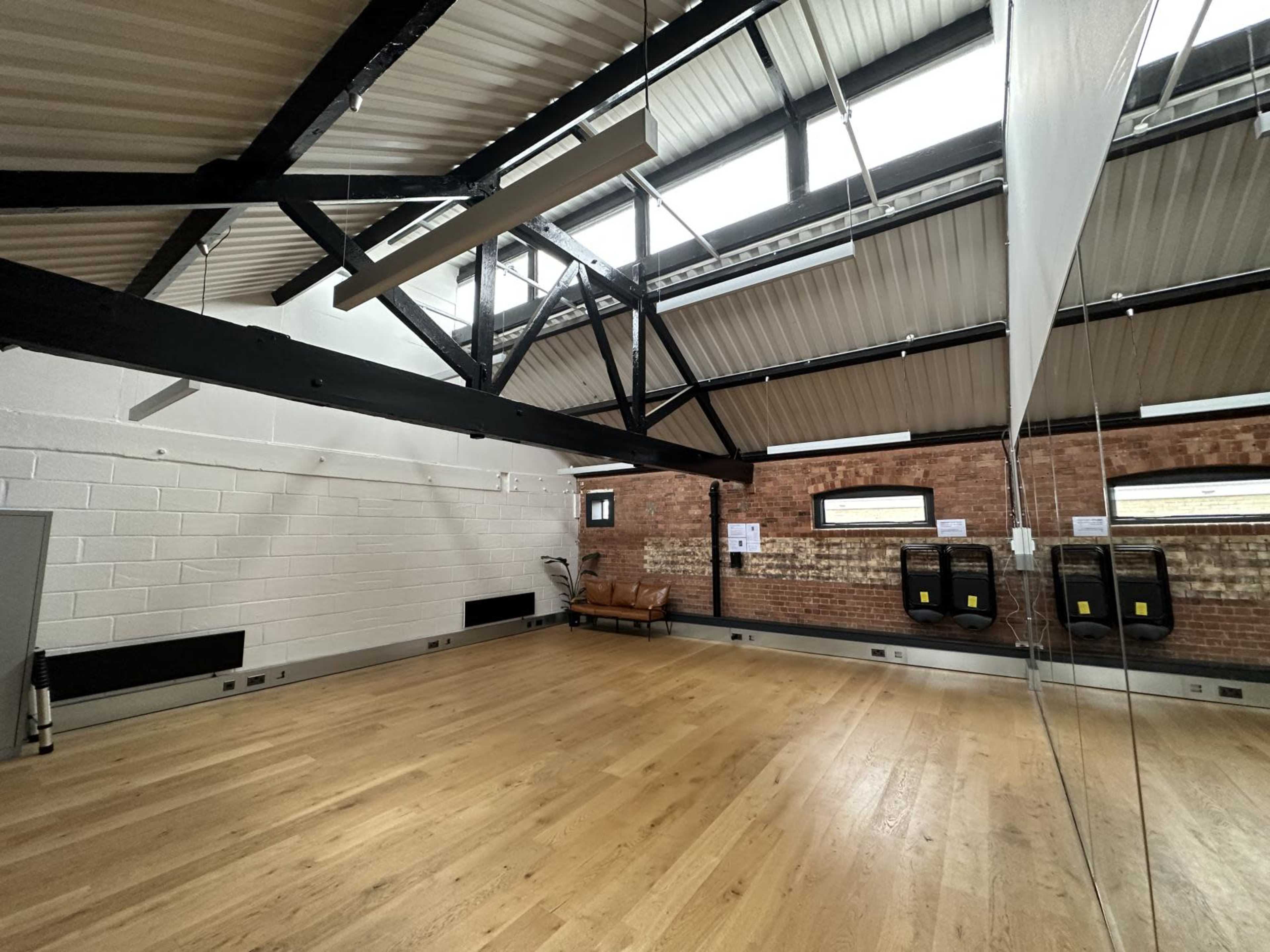The image shows an empty room with wooden flooring, exposed beams, large windows, and mirrors along one wall.