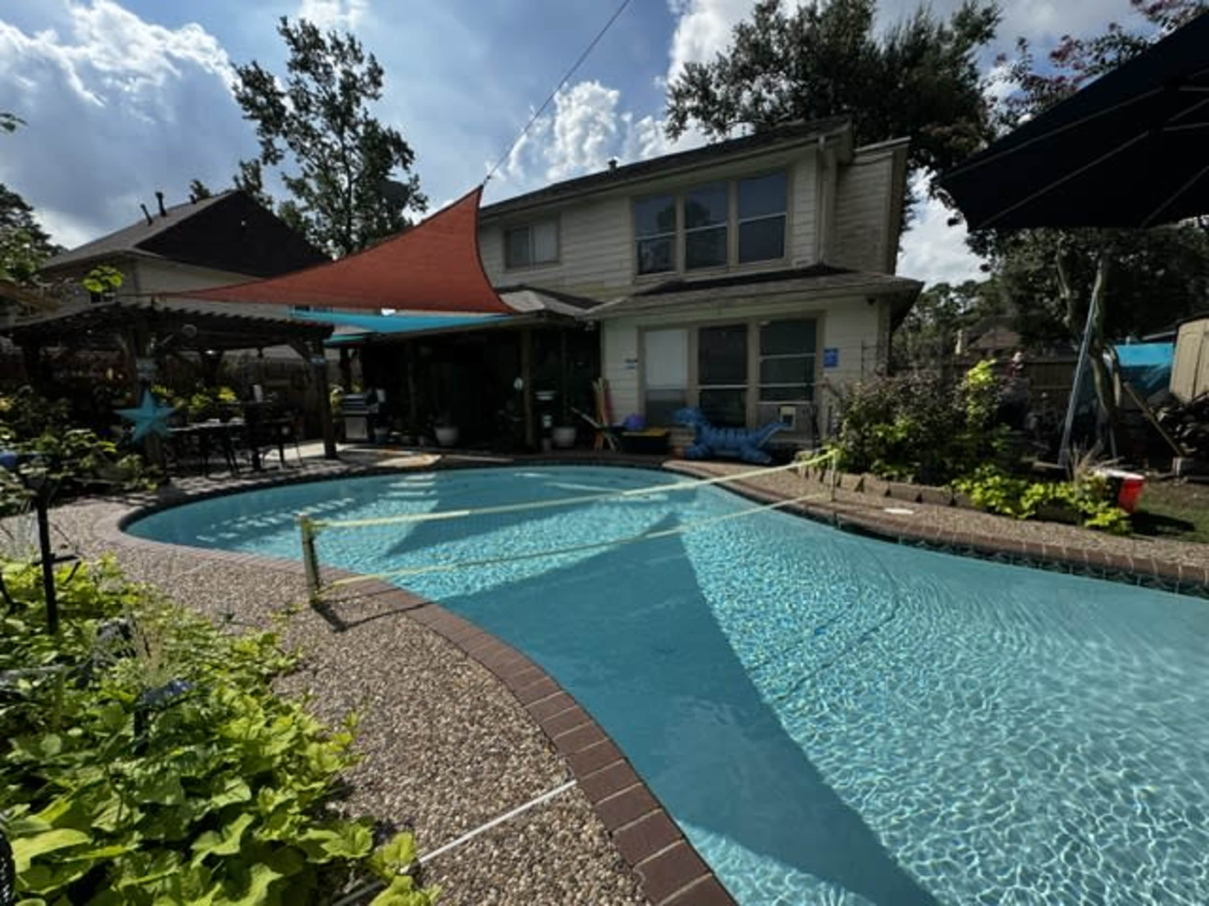 The image shows a backyard pool with a landscaped area, surrounded by a two-story home and various outdoor furniture.