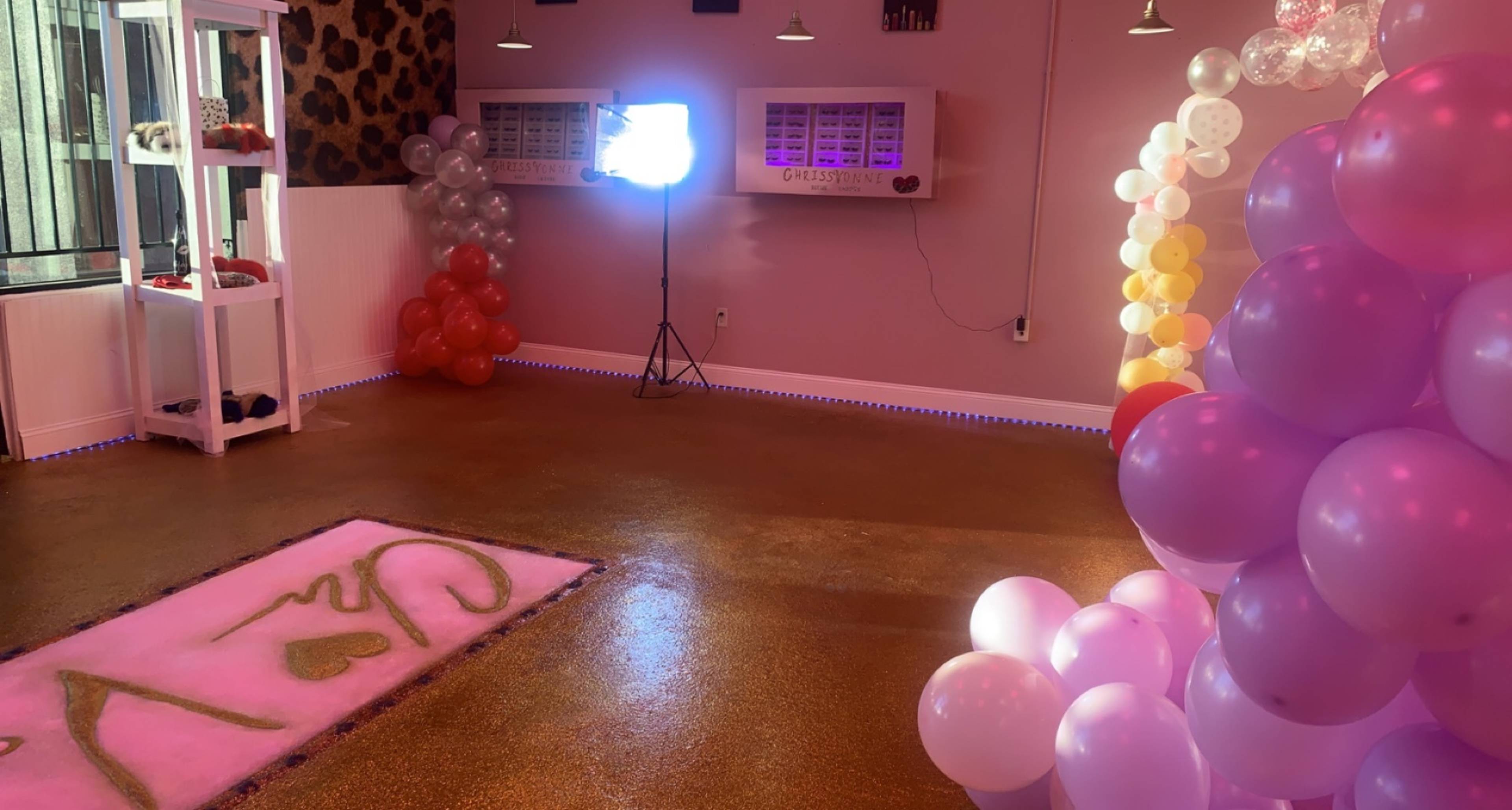 The image shows a brightly decorated room with colorful balloon arrangements, a pink carpet featuring the word "CAY," and a lighting setup in the corner.