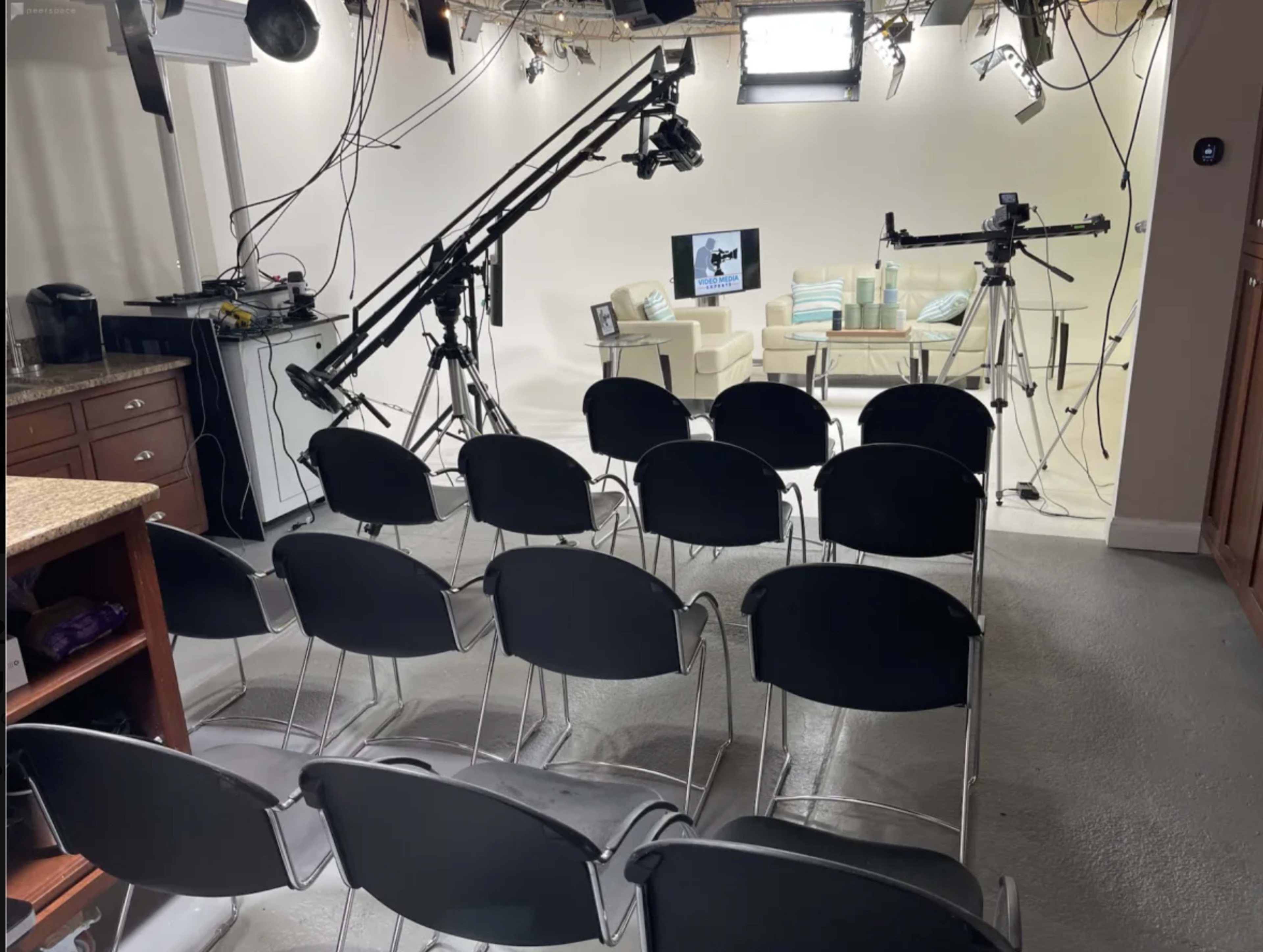A studio setup with several empty black chairs arranged in rows facing a set featuring a couch and cameras on tripods.