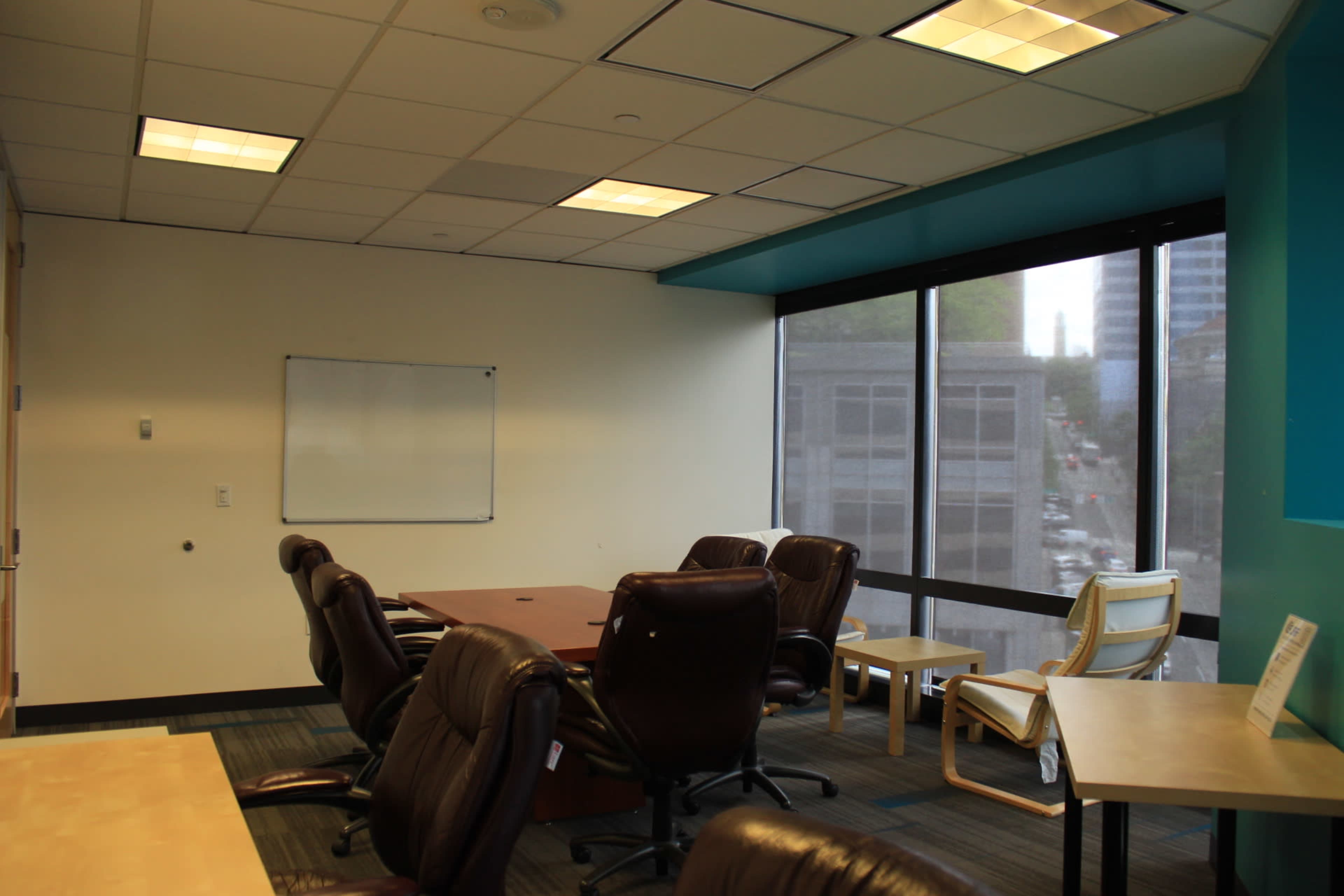 A meeting room with a wooden table, several office chairs, and a whiteboard, alongside a large window offering a view of an urban setting.