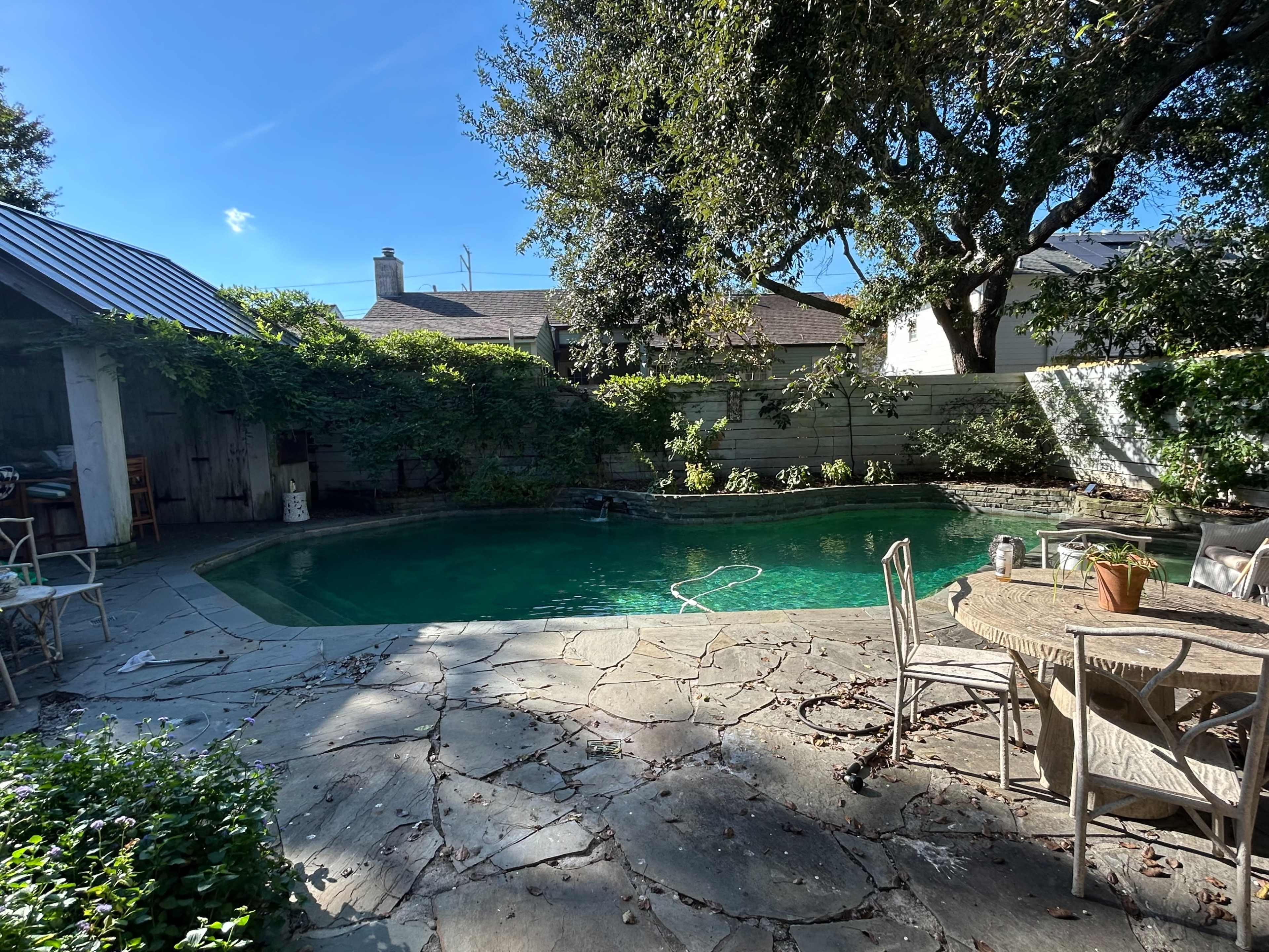 A small, partially shaded backyard pool area features a stone patio, surrounded by greenery and outdoor furniture.