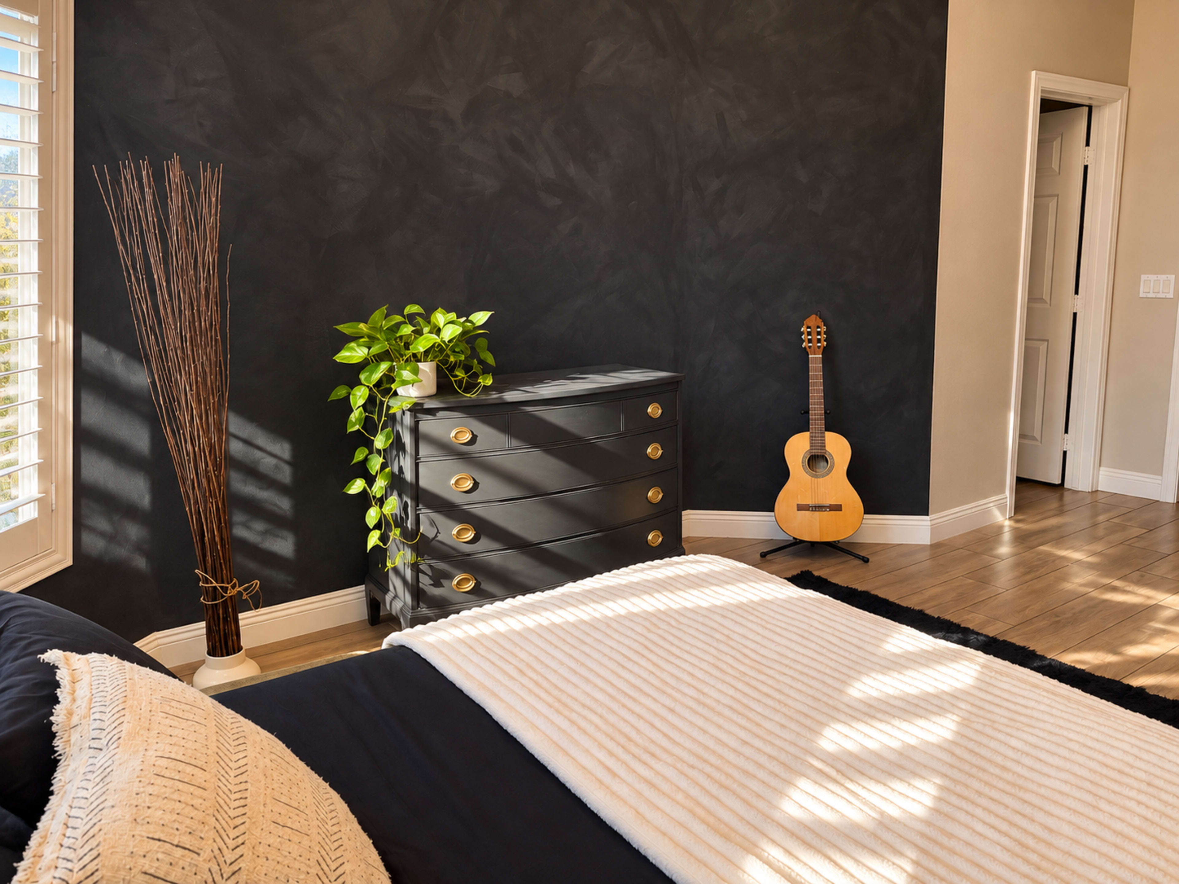 A modern bedroom features a dark accent wall, a tall plant next to a dresser, and an acoustic guitar leaning against the wall.