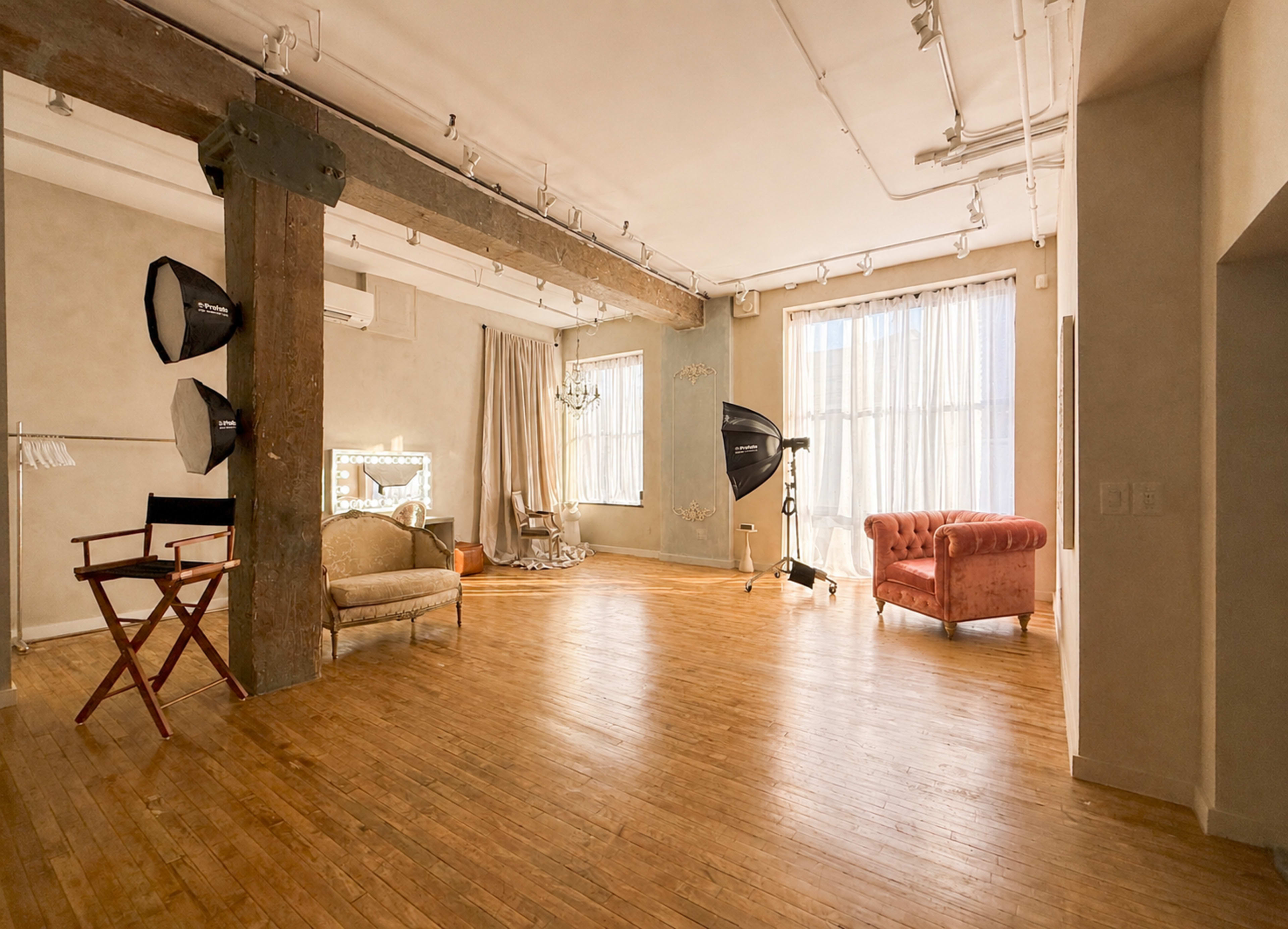 The image shows a spacious studio with wooden floors, large windows, and professional lighting equipment set up in a well-lit environment.