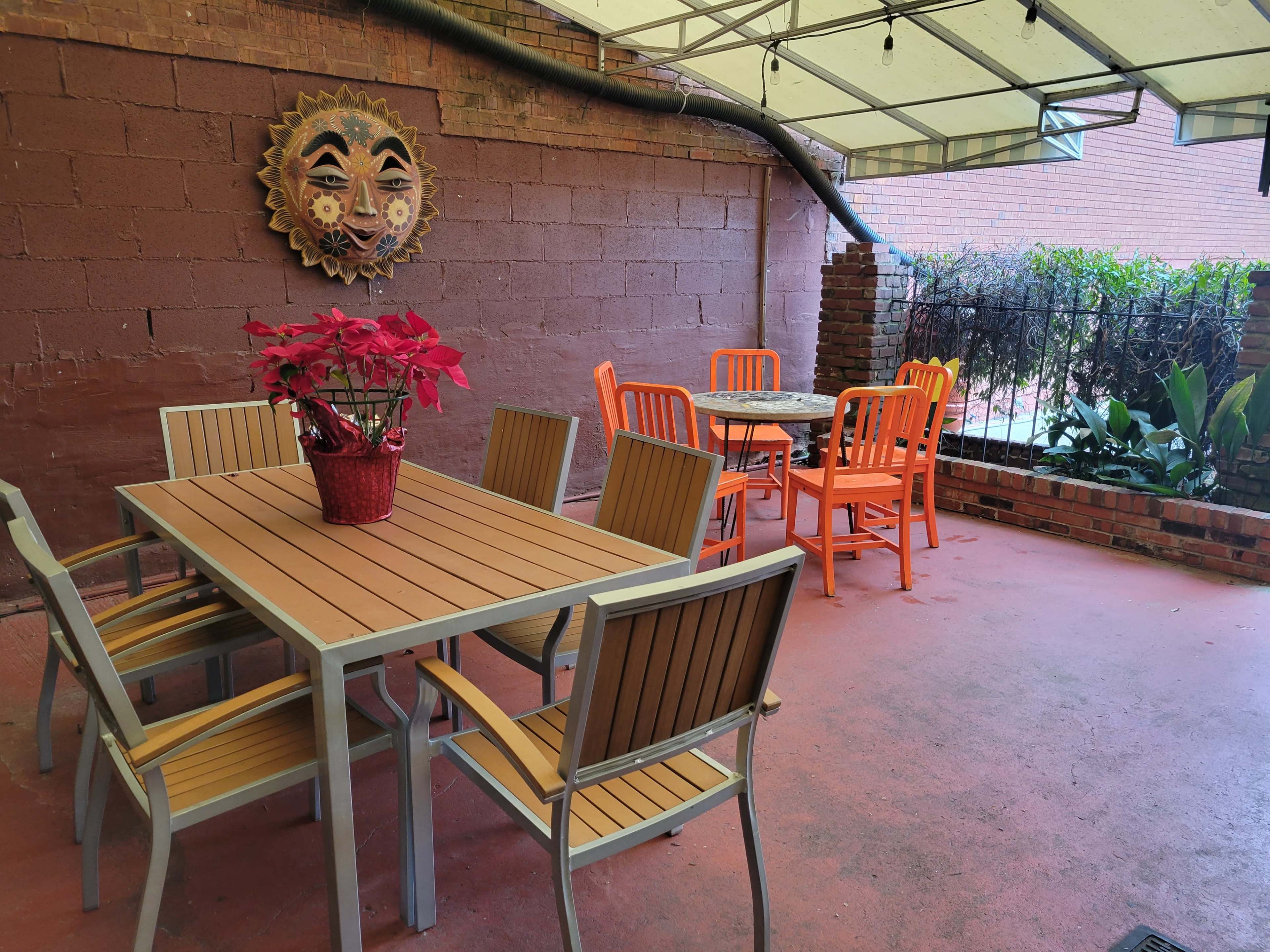 An outdoor seating area with a table and chairs under a canopy, featuring a decorative sun mask on the wall and a potted plant in the center.