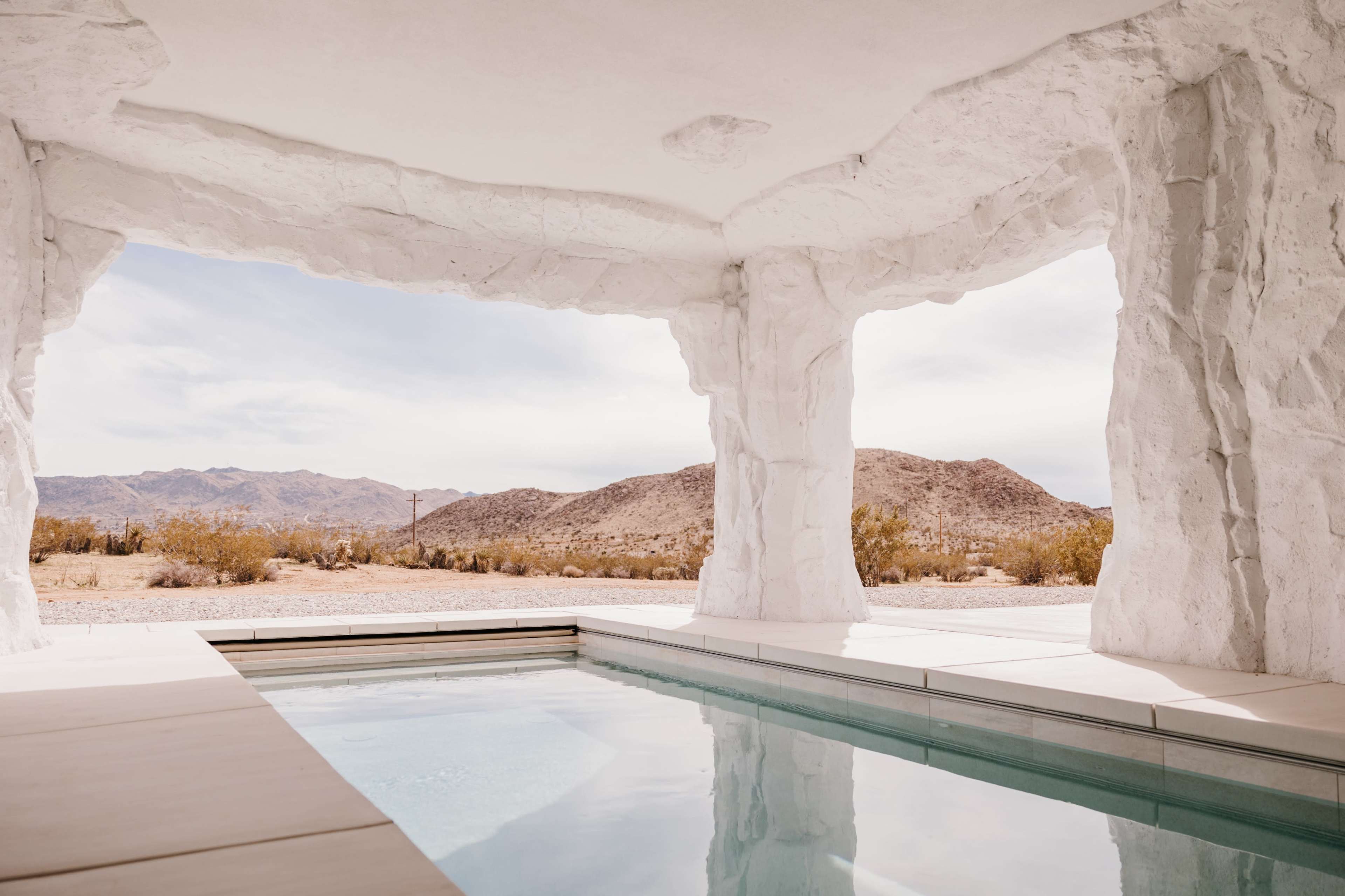 A modern, white-walled interior with a pool that opens up to a desert landscape framed by rock-like structures.