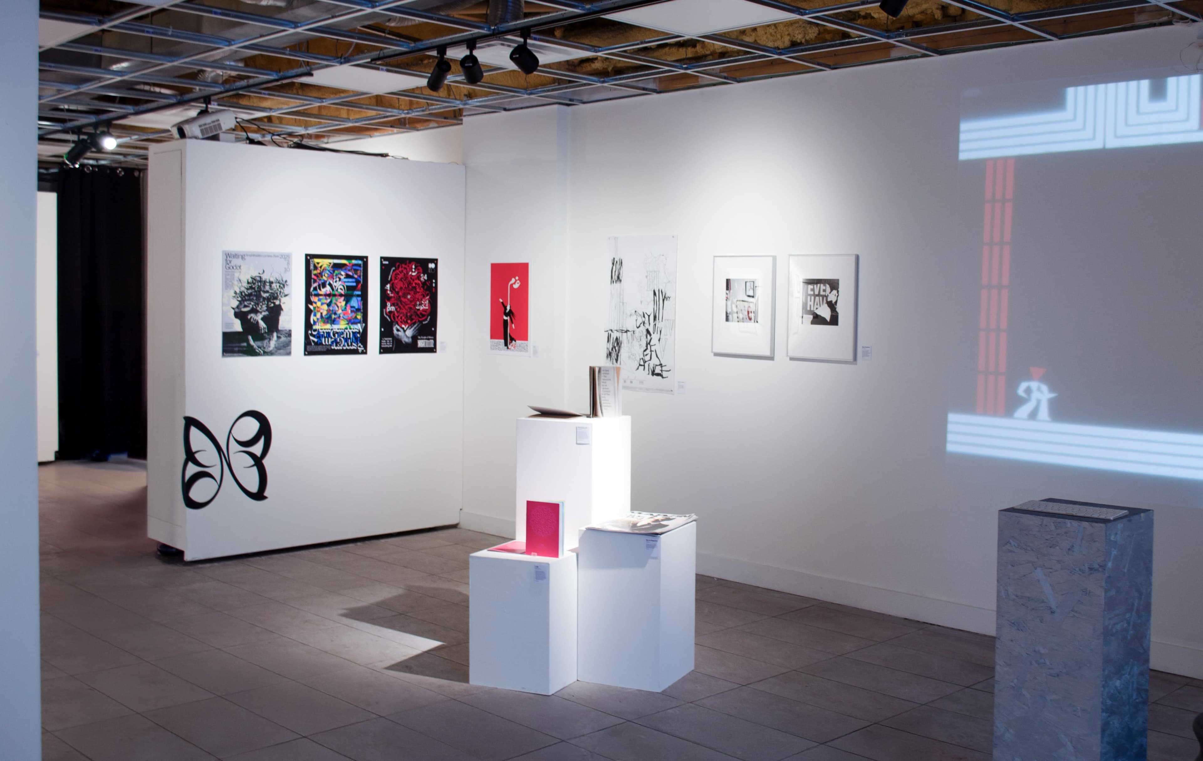 The image shows an art gallery interior with white walls displaying various framed artworks and a sculptural display stand in the center.