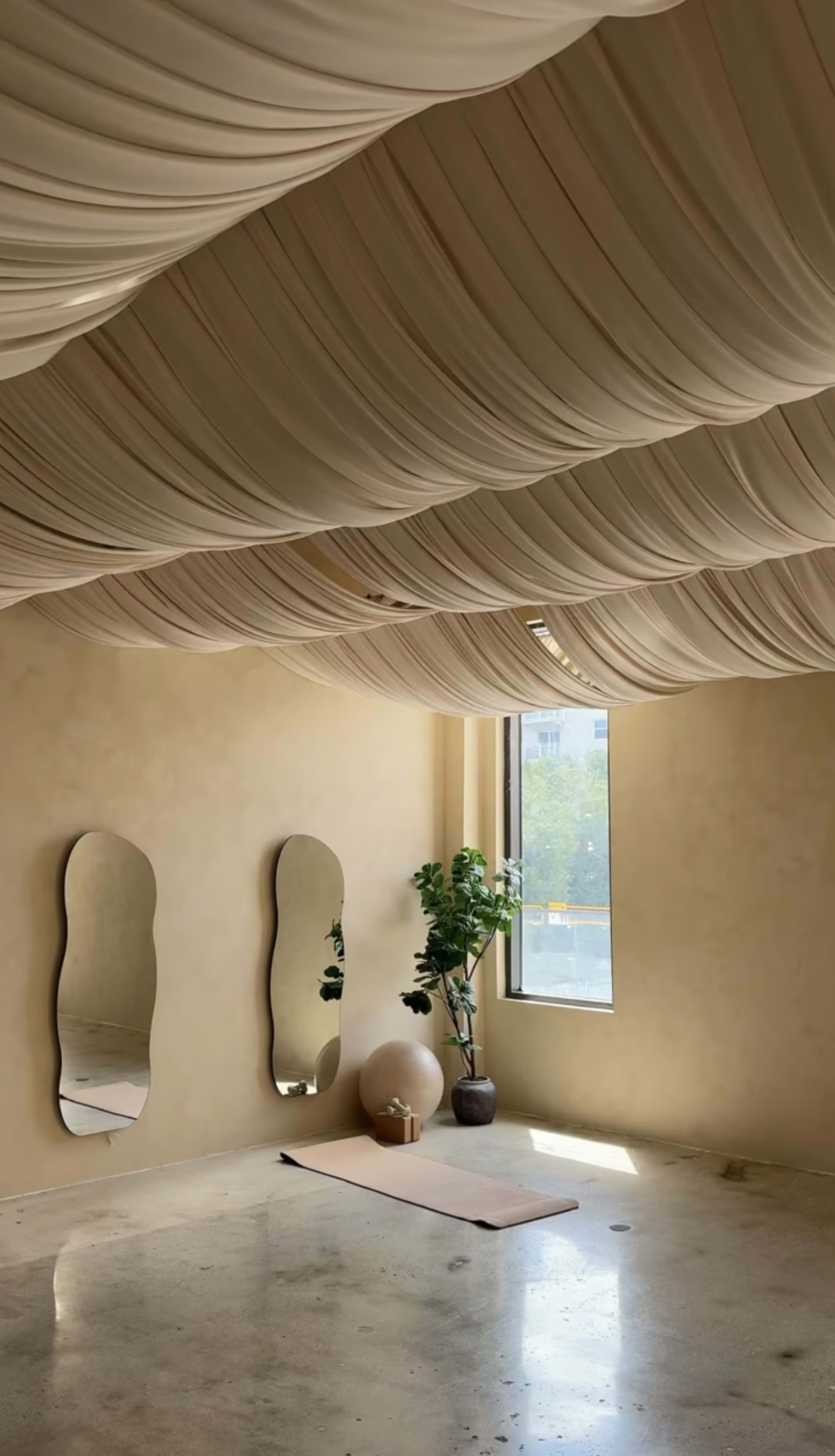The room features a soft, draped fabric ceiling, two mirrors on the walls, a potted plant, an exercise ball, and a yoga mat on a polished floor.