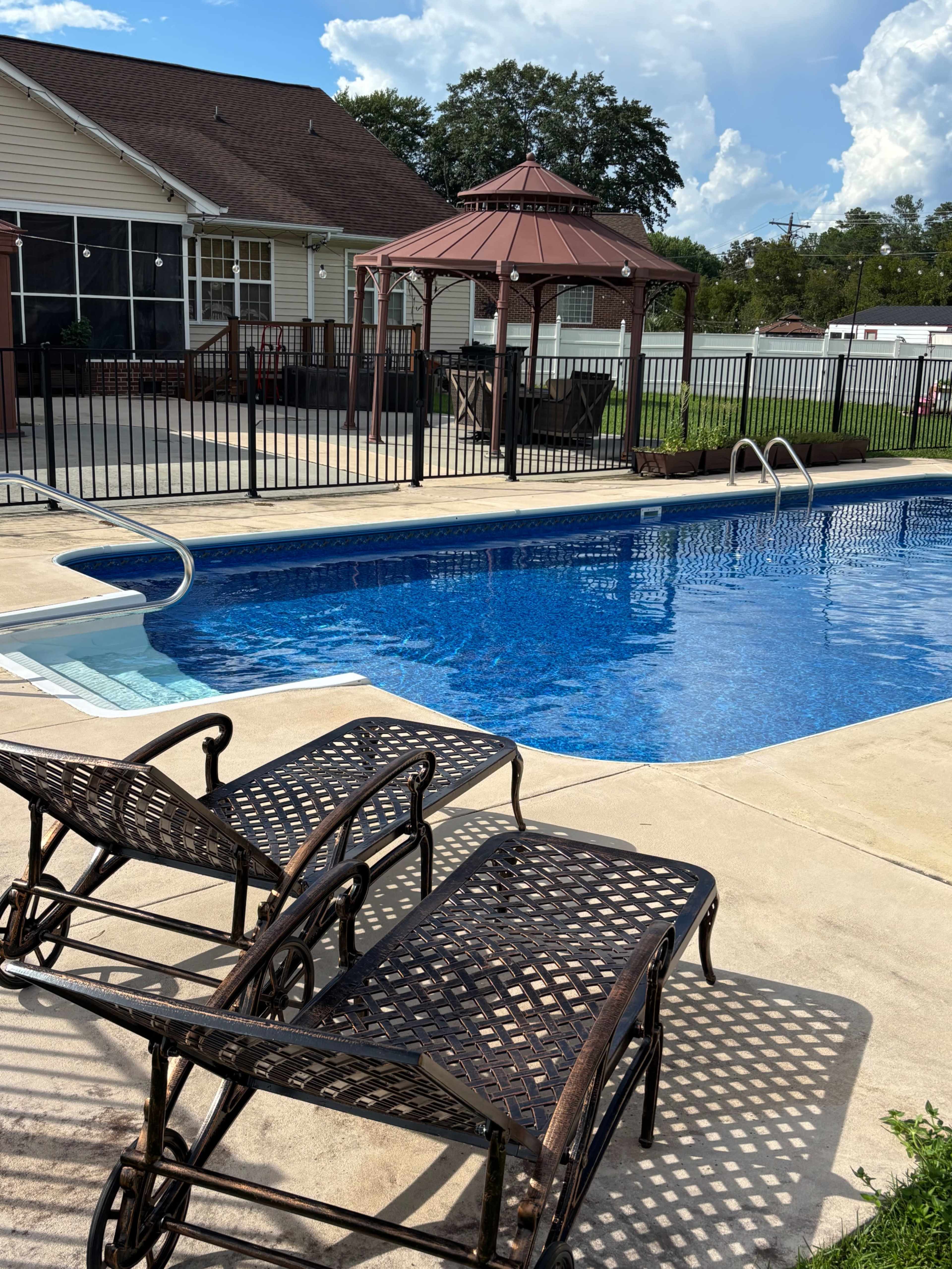 The image shows a swimming pool surrounded by a patio area with two lounge chairs and a gazebo in the background.