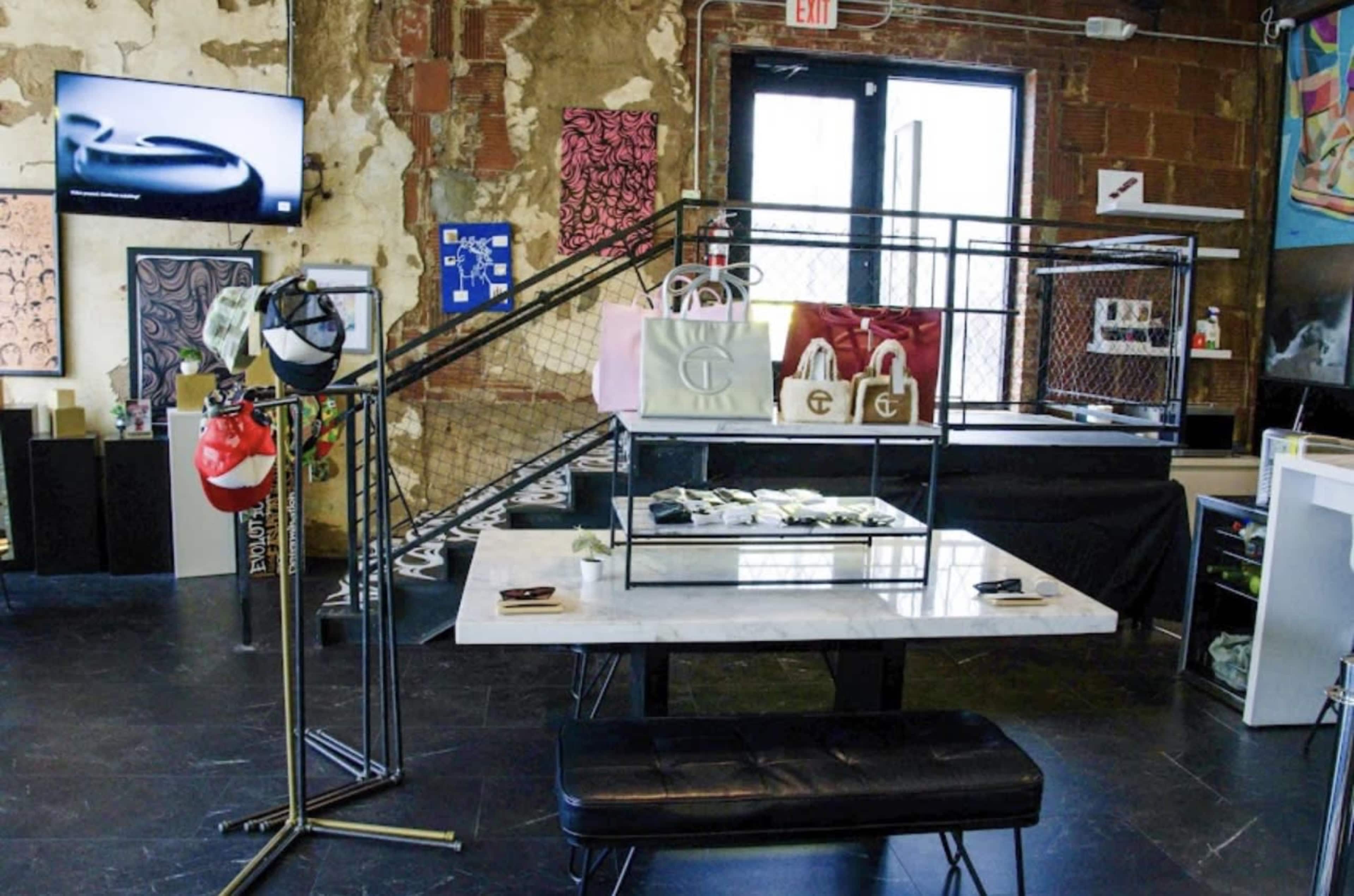The image shows a retail space featuring a display table with handbags, a black bench, and a wall with colorful artwork and a television screen.