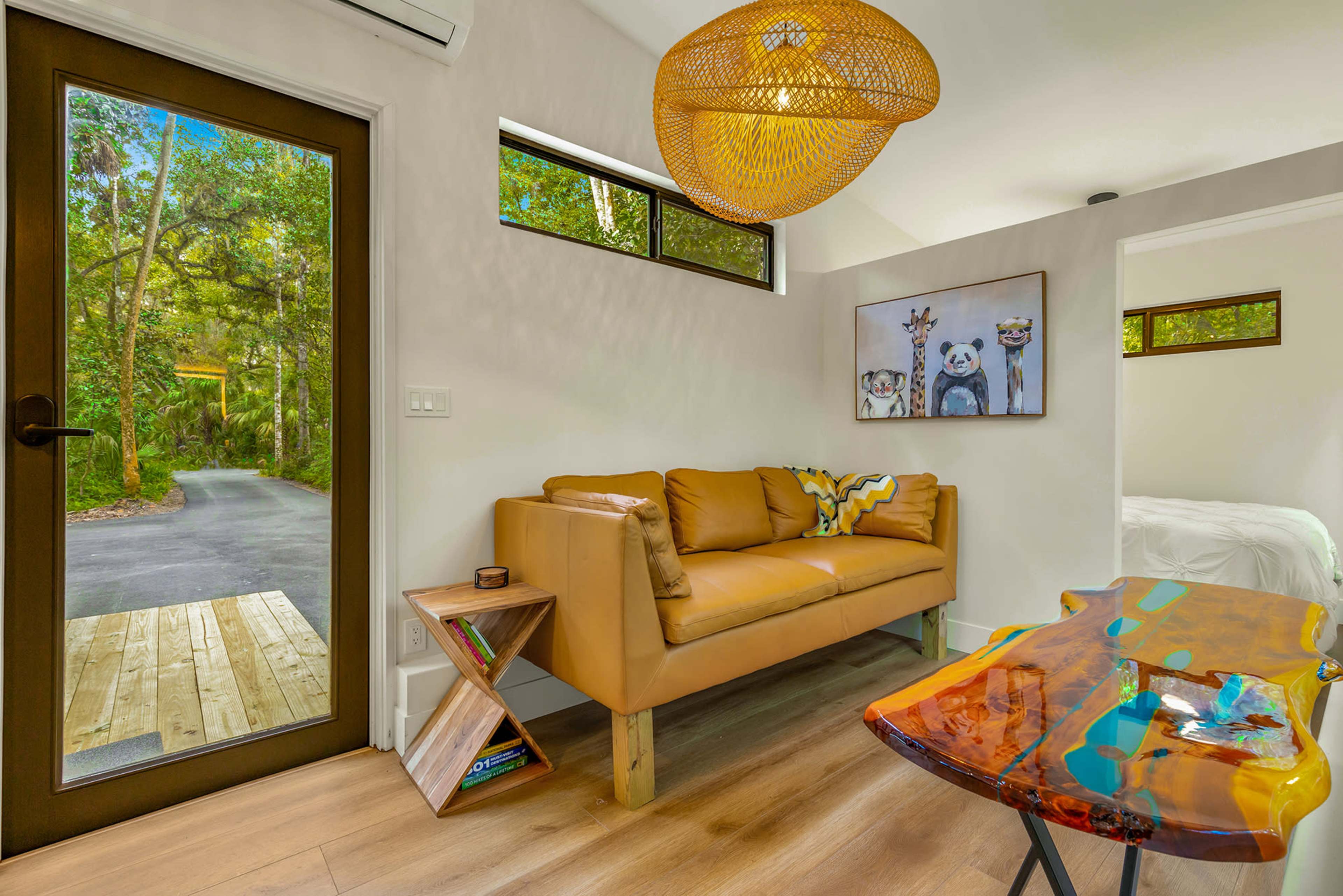 A cozy interior features a brown sofa, a colorful coffee table, and artwork hanging on the wall, with a view of a tree-lined pathway visible through the open door.