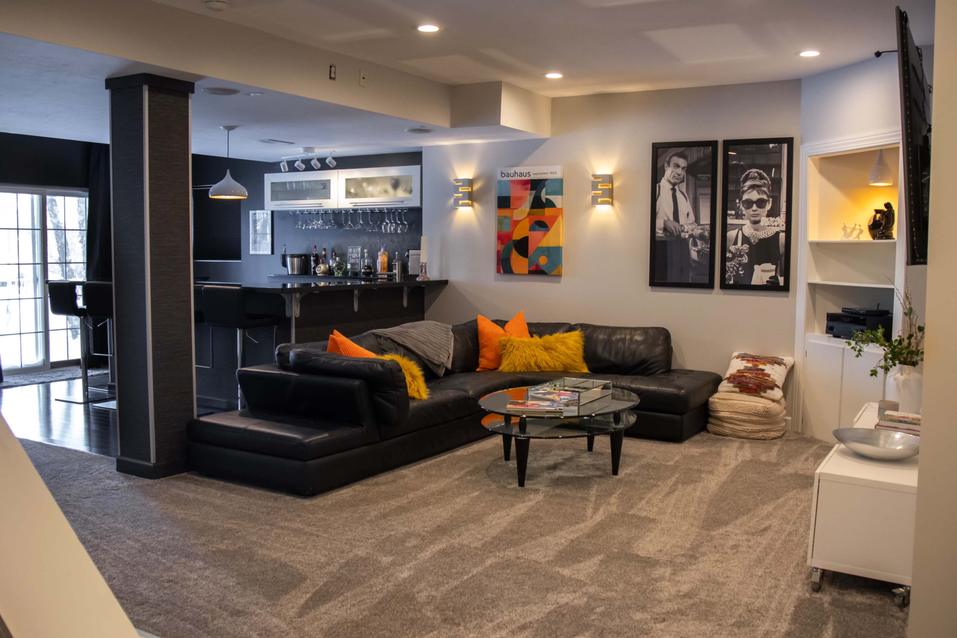 The image shows a modern living room with a sectional black couch, a glass coffee table, and a bar area in the background, complemented by artwork on the walls and warm lighting.