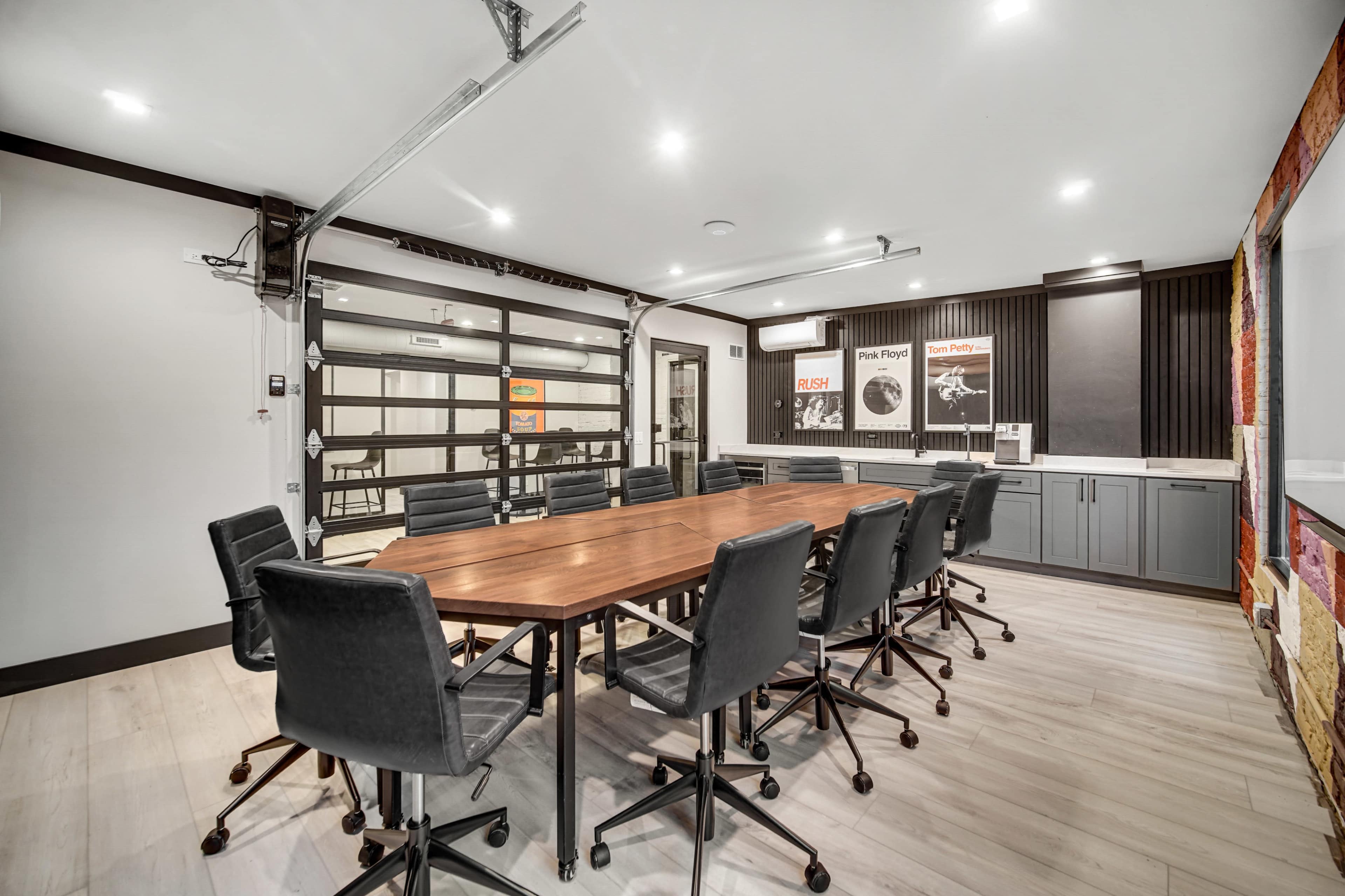 The image shows a modern conference room featuring a large rectangular wooden table surrounded by black rolling chairs, with a garage-style door and various wall posters visible.