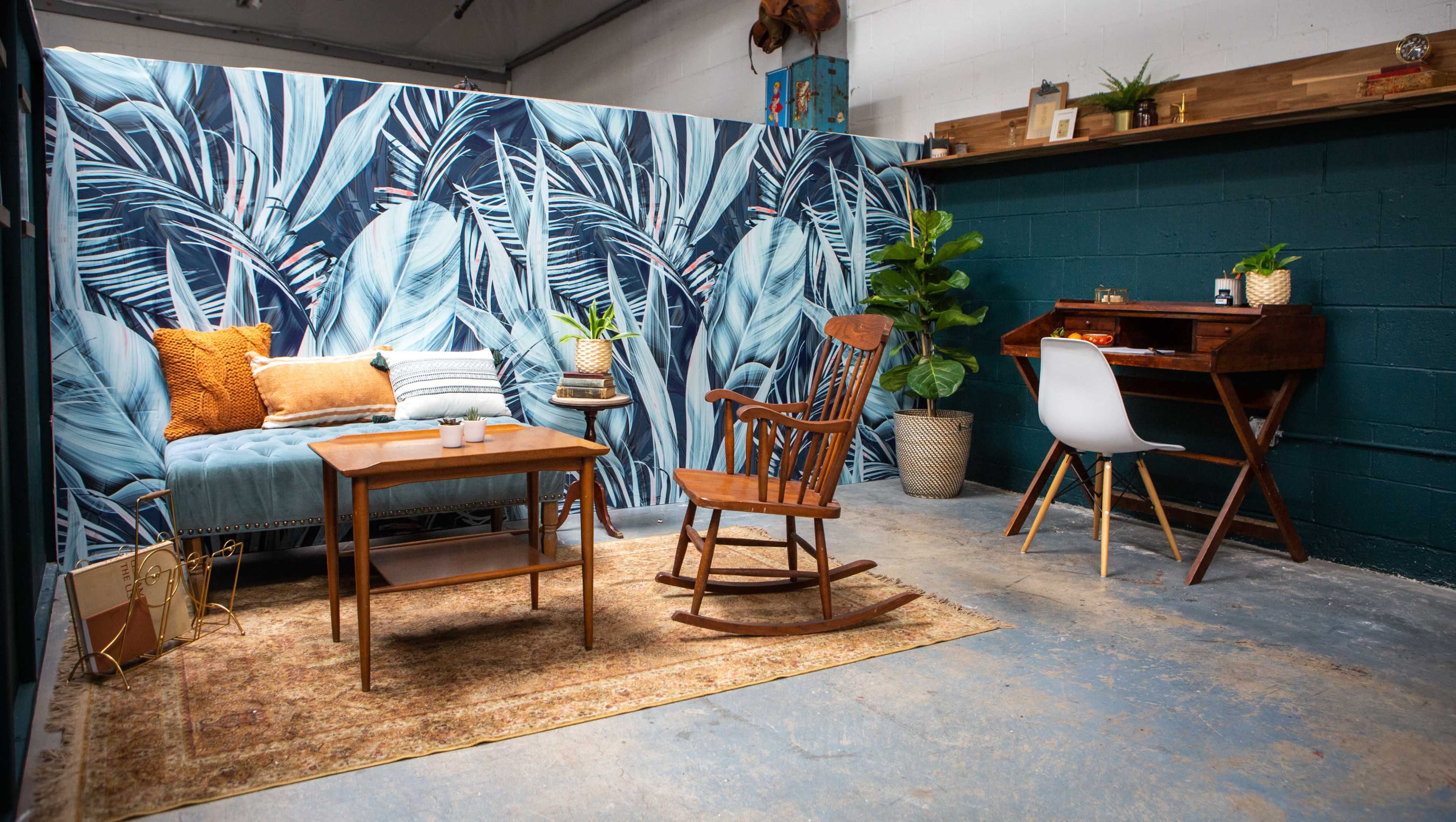 A cozy interior space featuring a wooden rocking chair, a small coffee table, and a writing desk against a backdrop of vibrant palm leaf wallpaper.