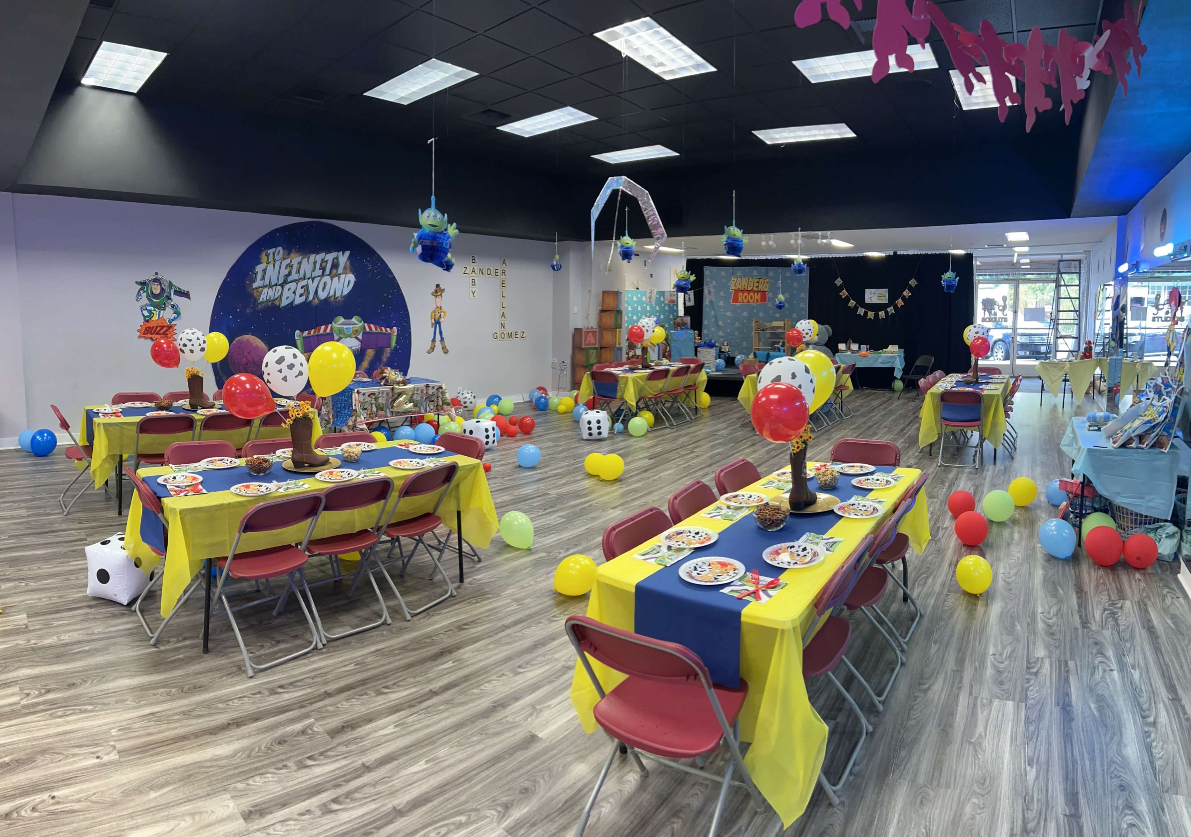 The scene shows a brightly decorated party venue with tables set for an event, featuring colorful tablecloths, balloons, and themed decorations.