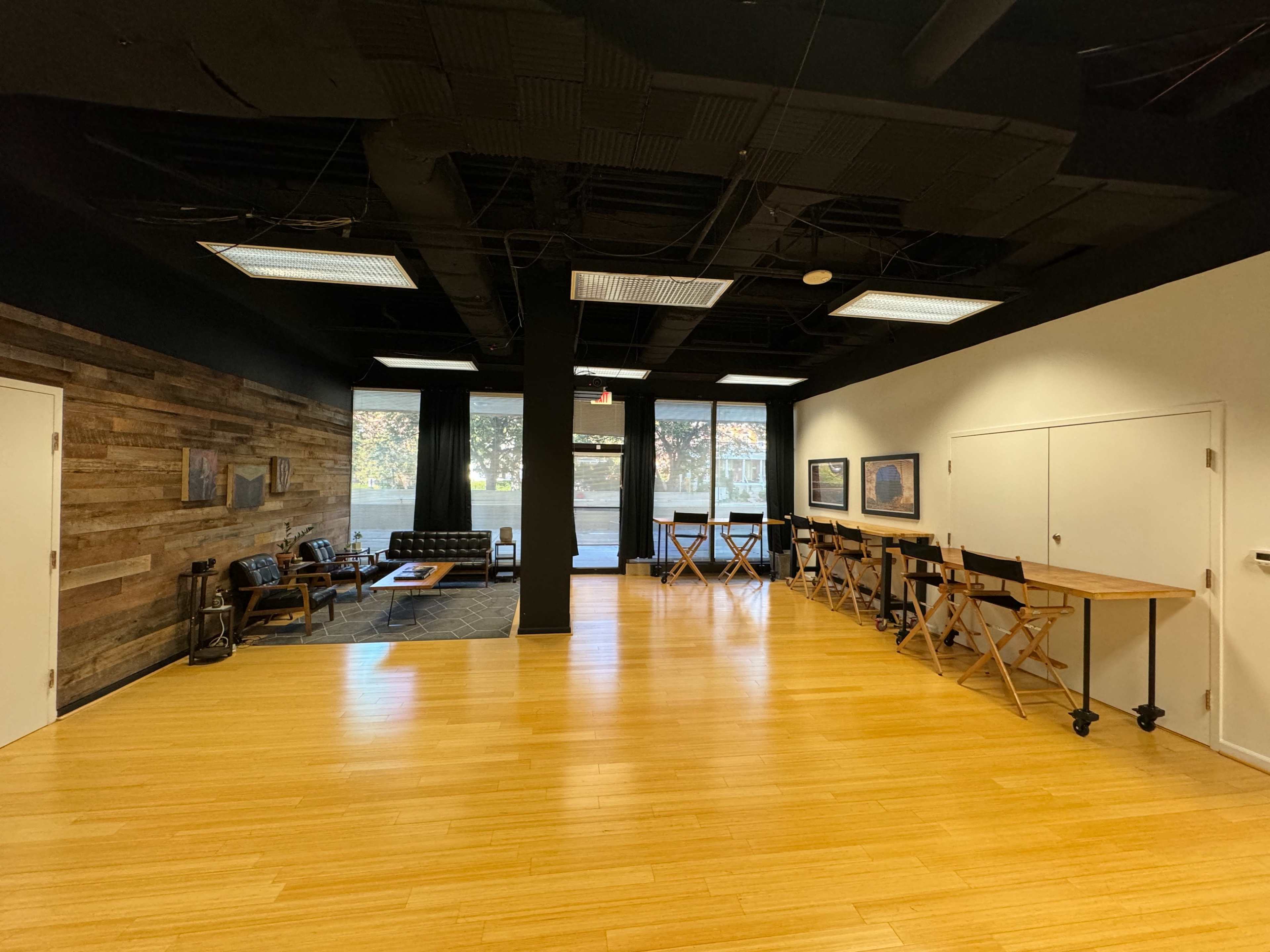 The image shows a spacious, modern room with wooden flooring, a seating area on the left, and a long table with director's chairs on the right.