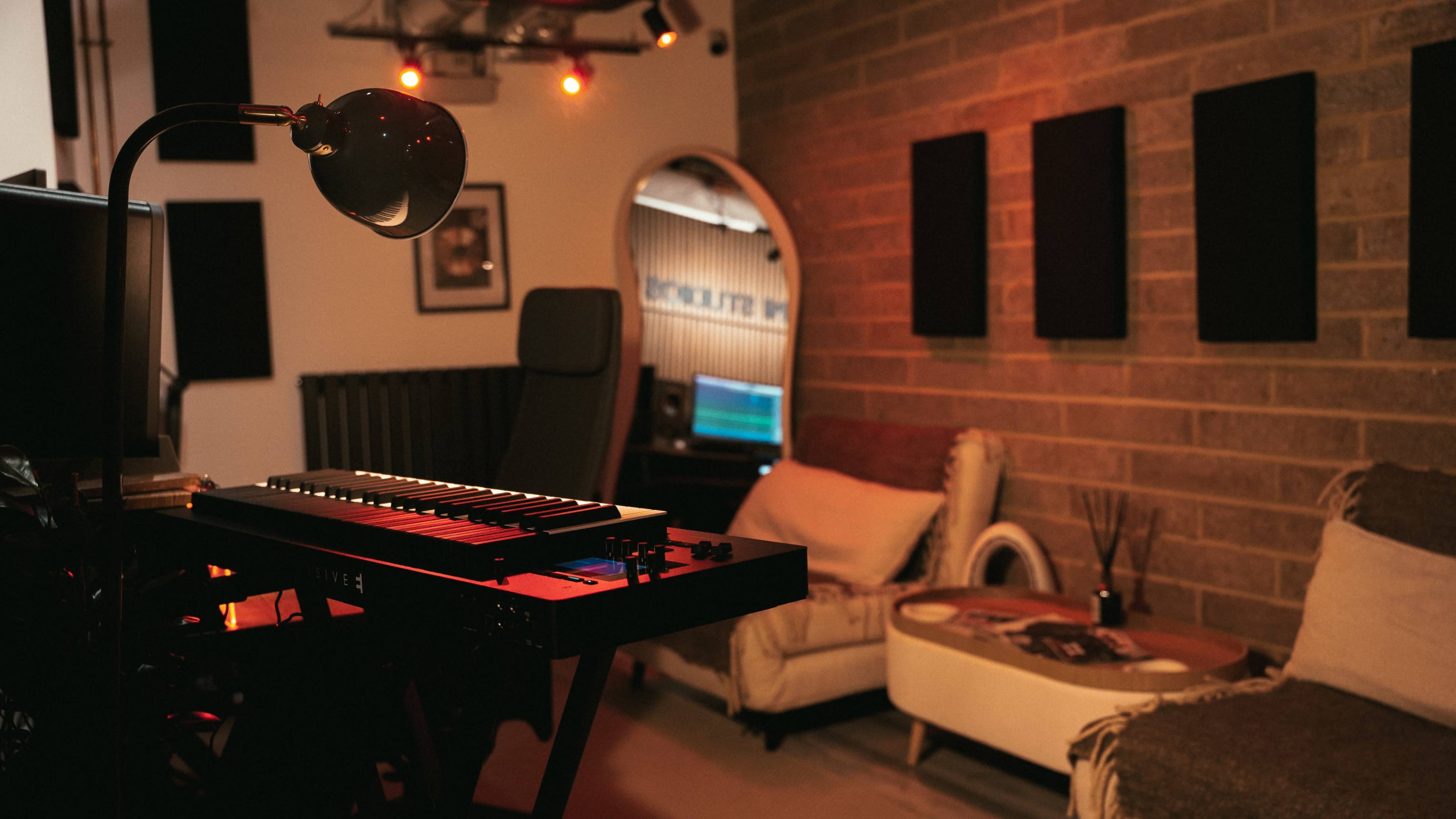 The image shows a cozy music studio with a synthesizer and a comfortable seating area featuring two armchairs and a coffee table, against a backdrop of exposed brick walls and acoustic panels.