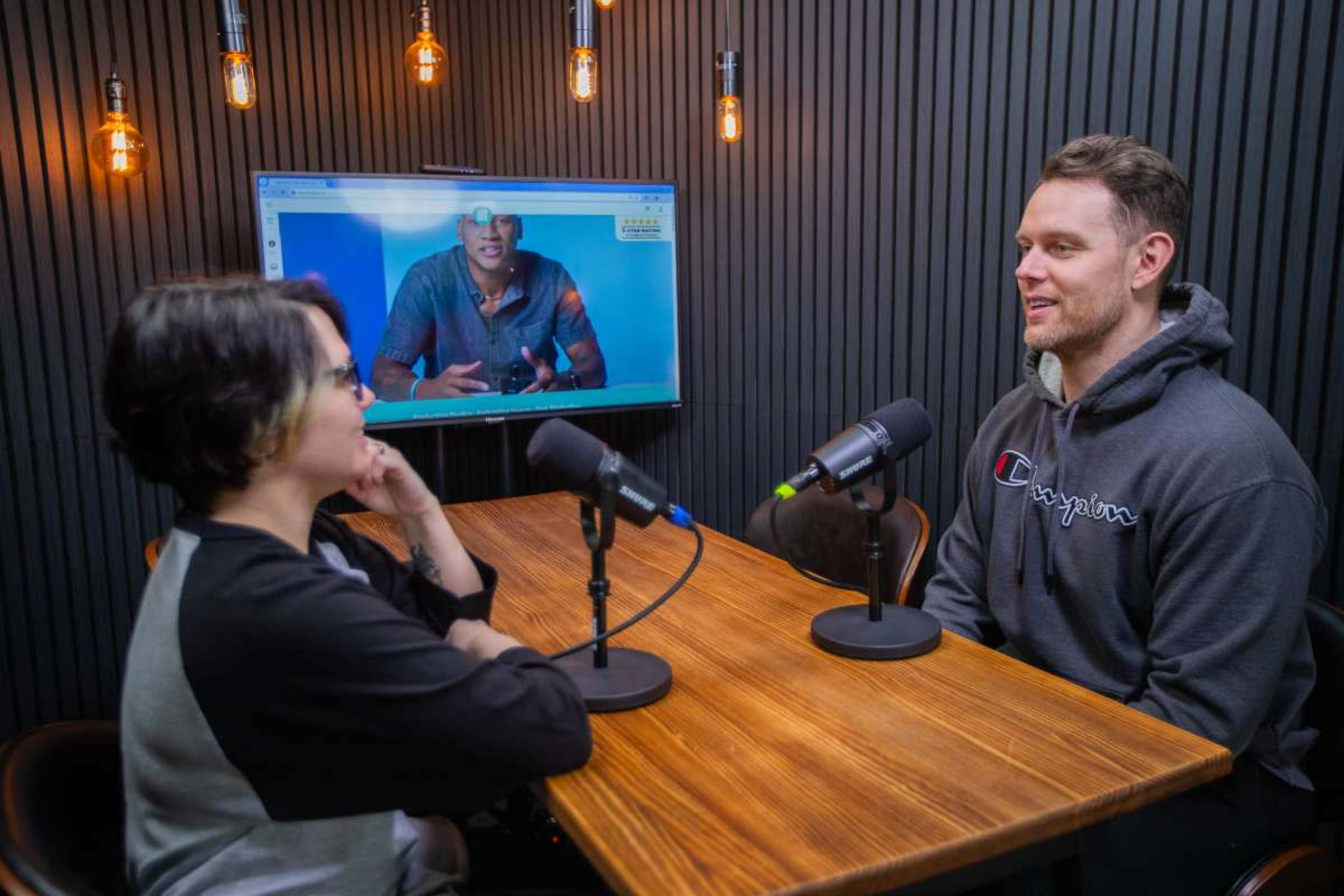 A man and a woman sit across from each other at a table with microphones, while a large screen in the background displays a video of another man.