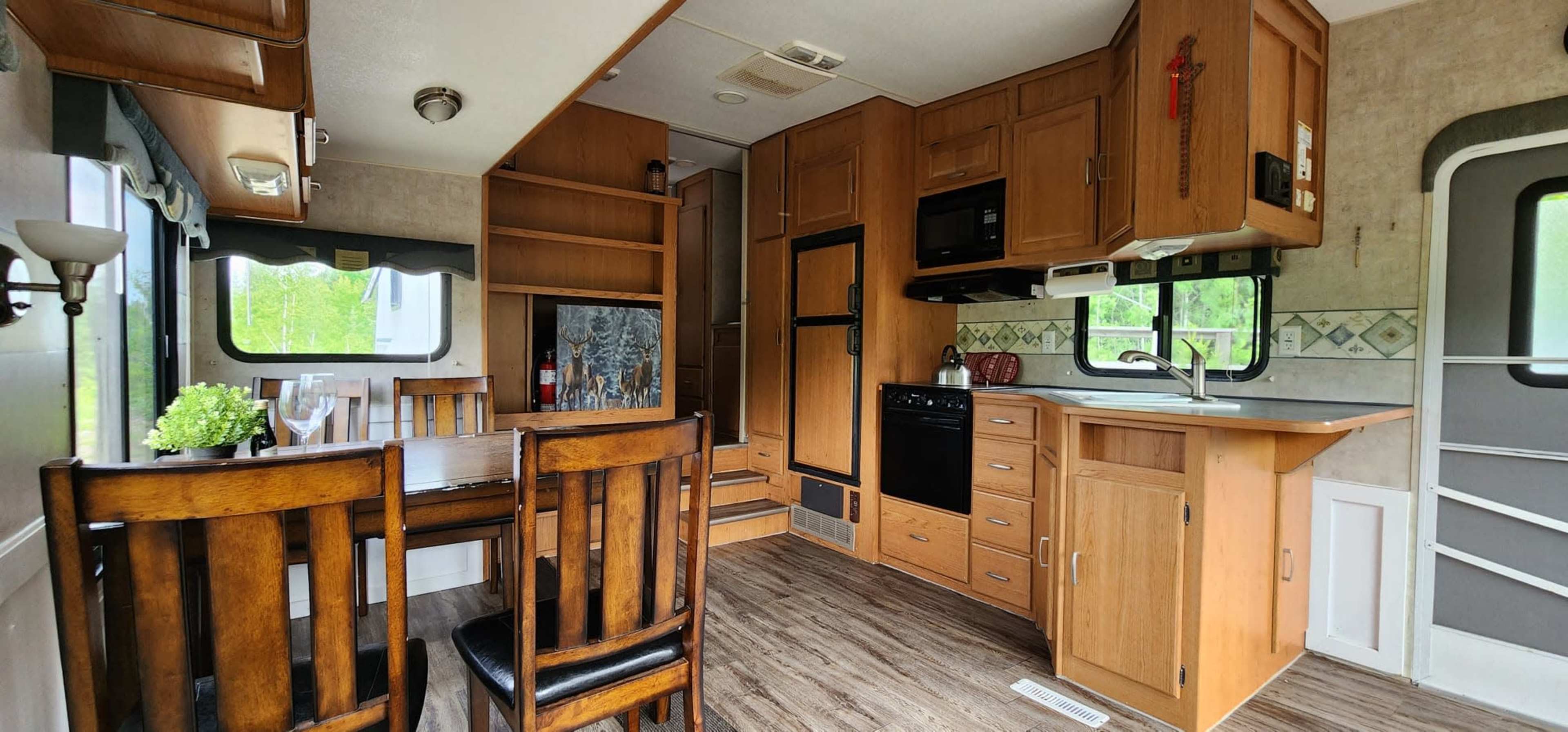 The interior of a camper showcases a wooden dining table and chairs, a kitchen area with appliances, and cabinetry, all set against a backdrop of windows.