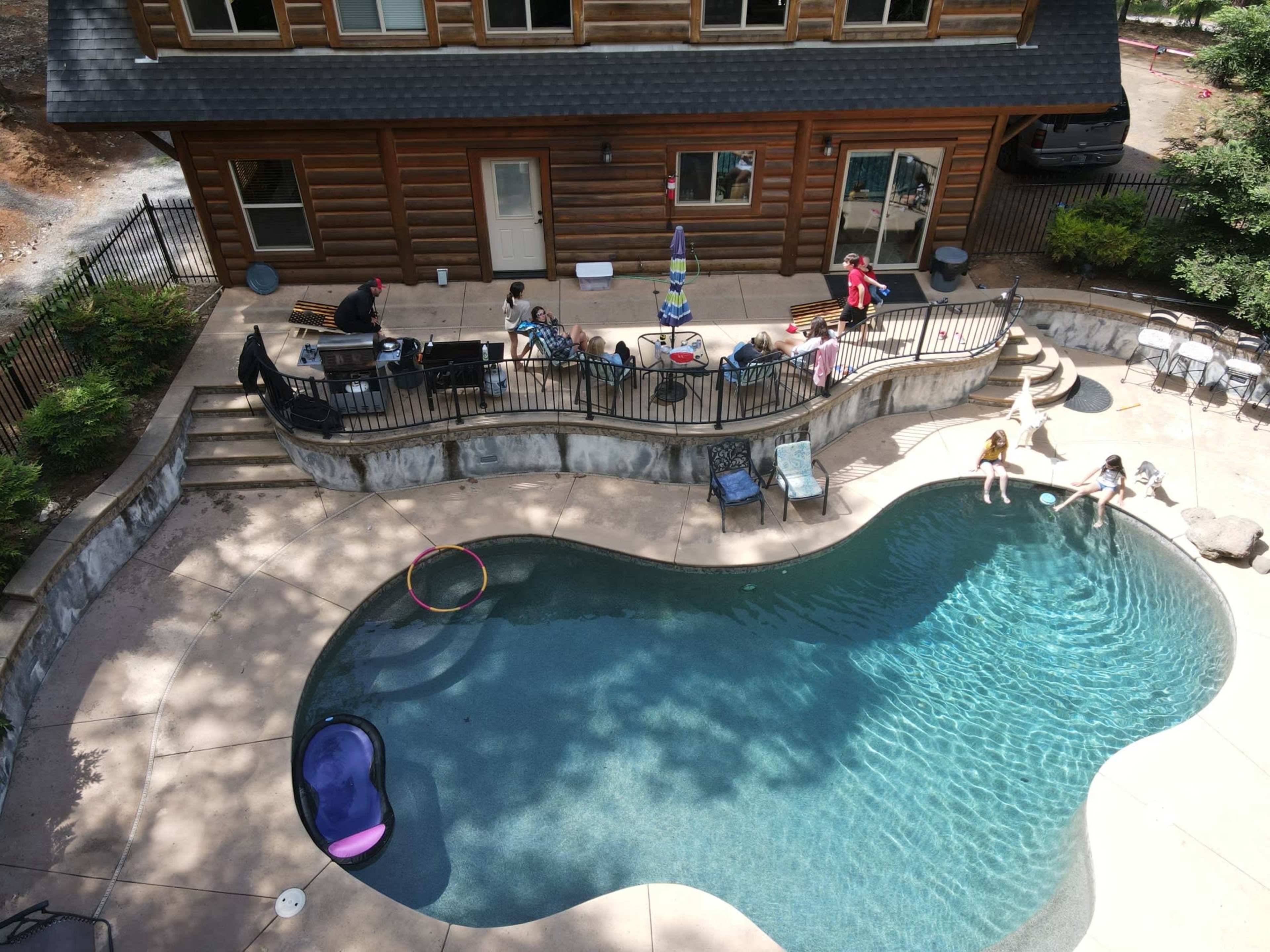 A group of people gather around a swimming pool next to a log cabin, enjoying the outdoor space with lounge chairs and patio furniture.