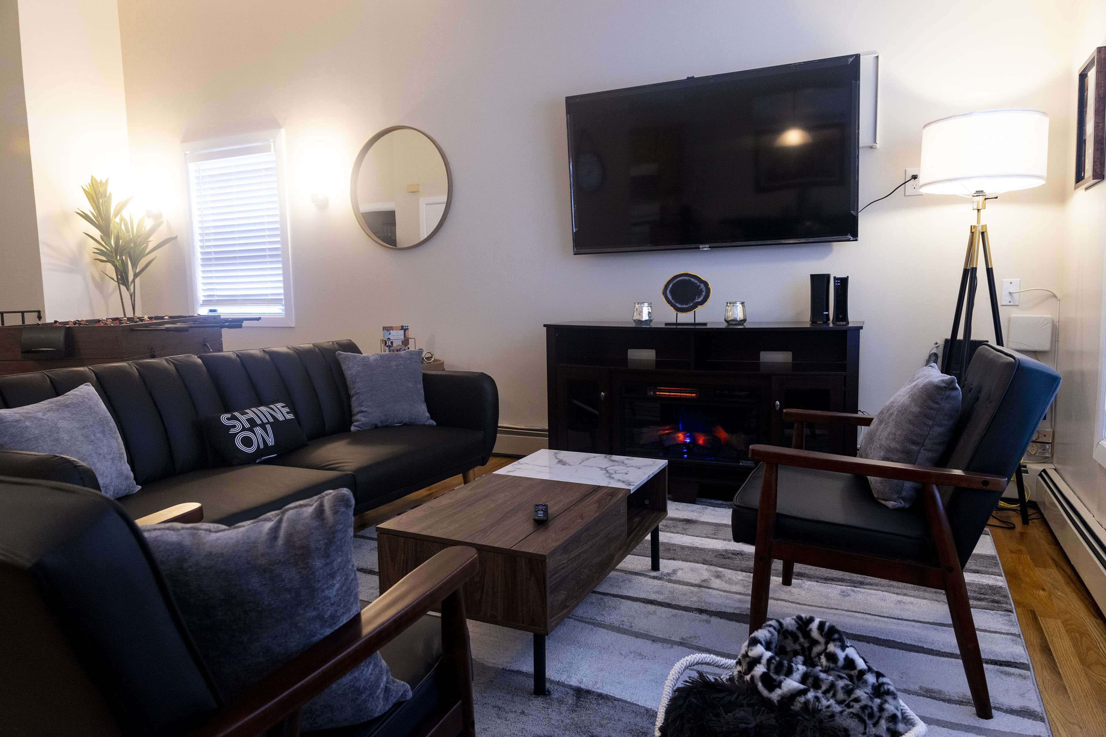 The living room features a black sofa with decorative pillows, a wooden coffee table, an electric fireplace, and a large television mounted on the wall.