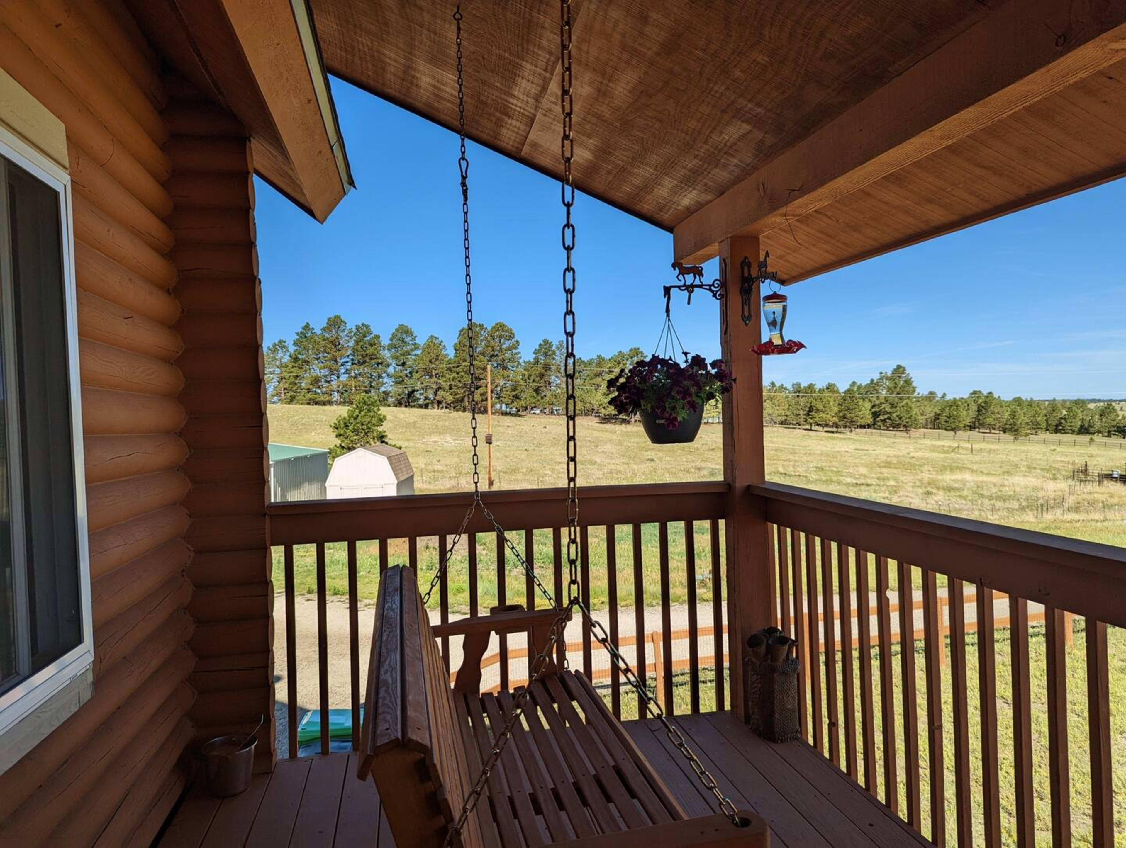A wooden porch swing hangs from a chain on a log cabin balcony, overlooking a grassy field and trees under a clear blue sky.