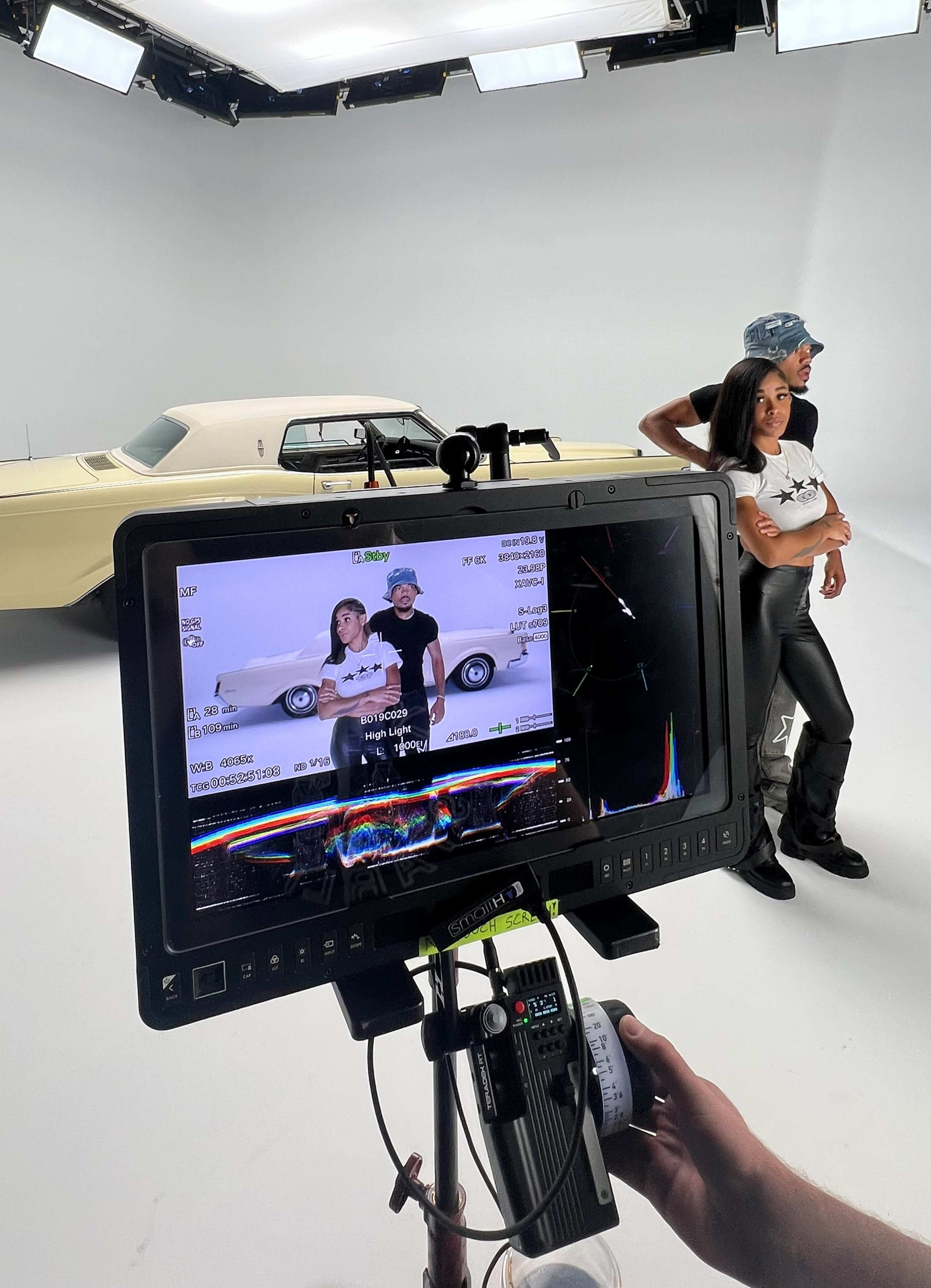 A film crew sets up a shot featuring two individuals posing in front of a classic car against a plain backdrop.