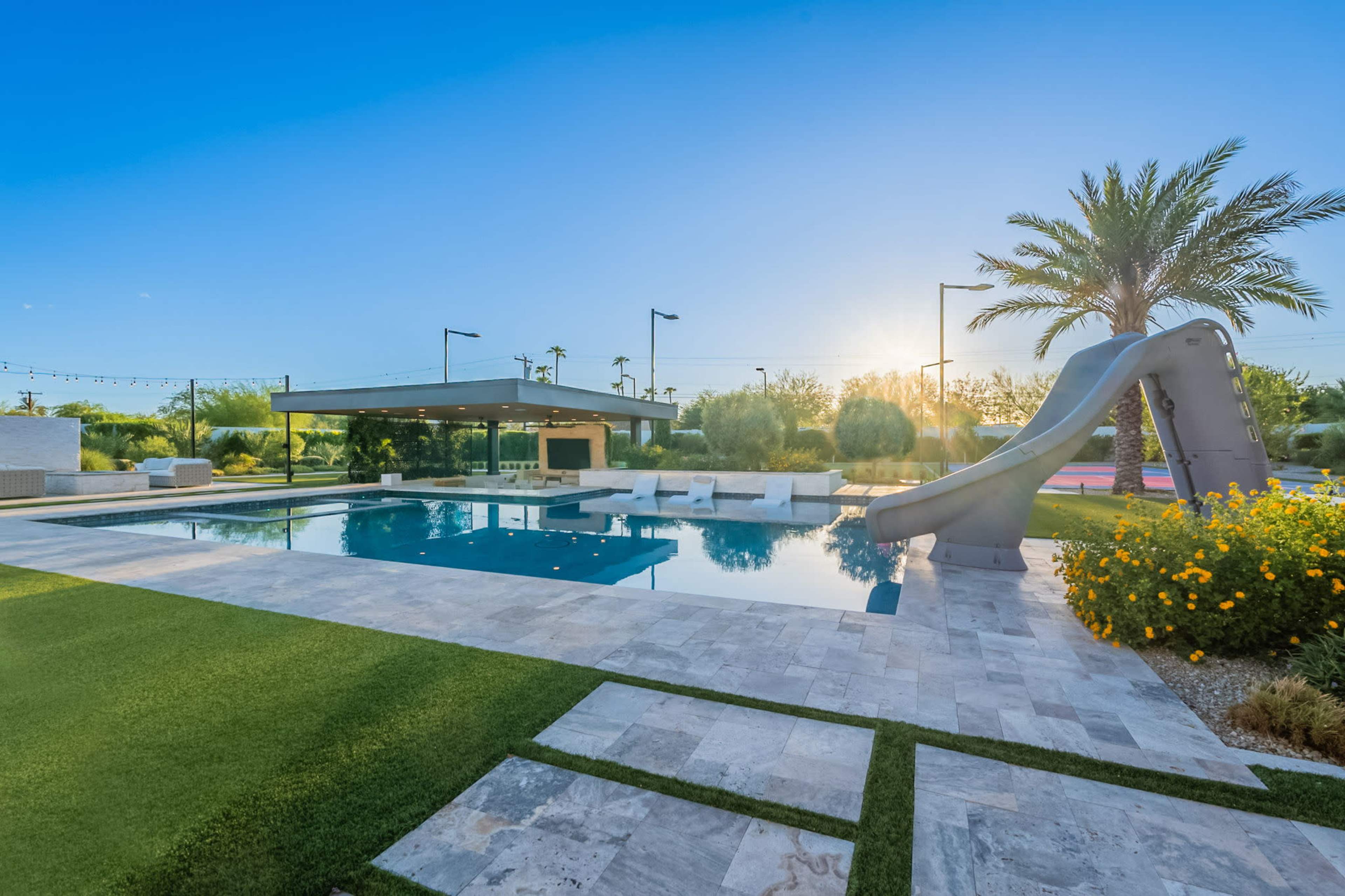 The image shows a modern outdoor pool area featuring a large swimming pool with a slide, surrounded by landscaped greenery and seating spaces.