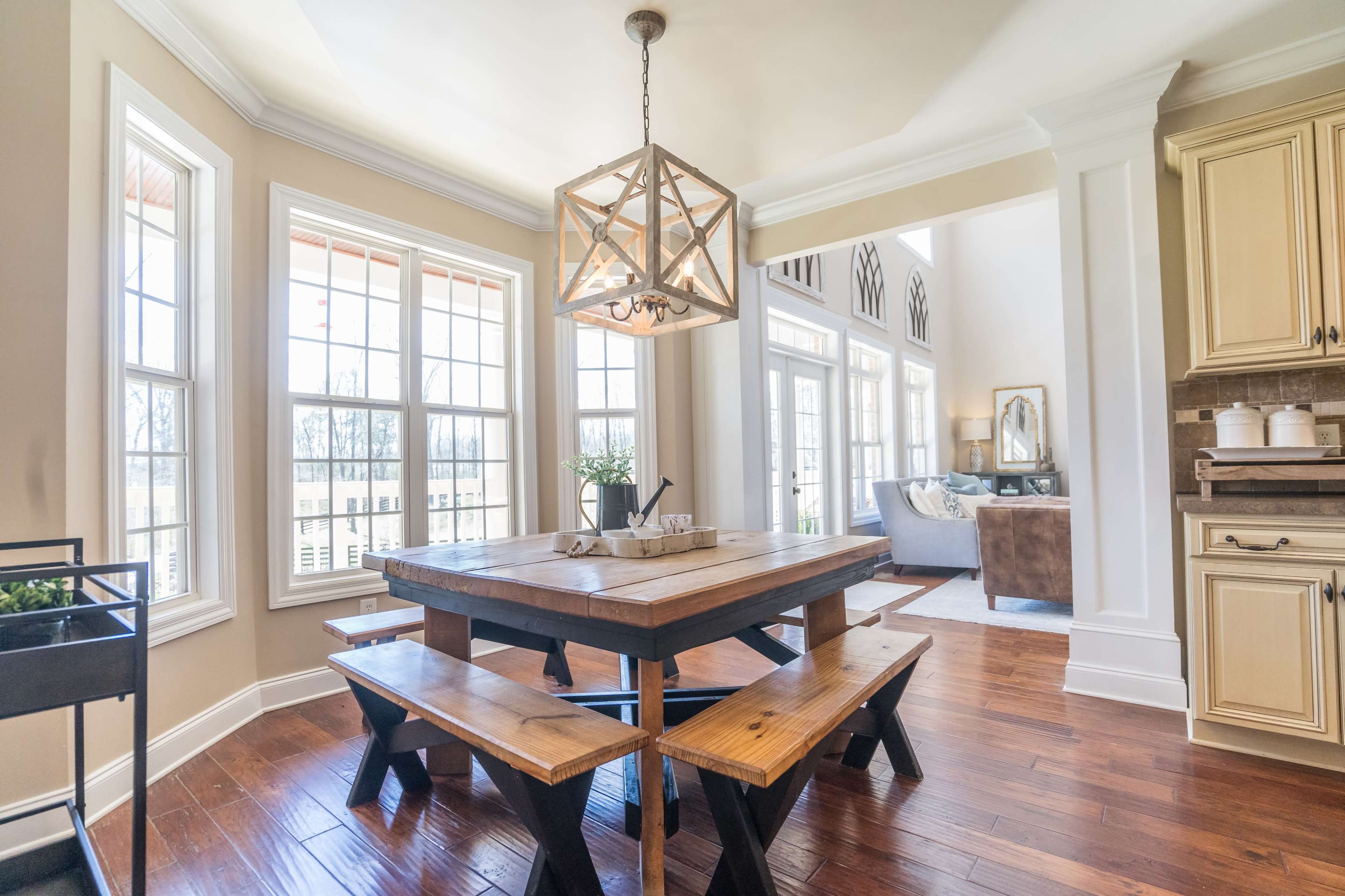 A wooden dining table with bench seating is positioned under a geometric chandelier in a bright, open dining area with large windows.