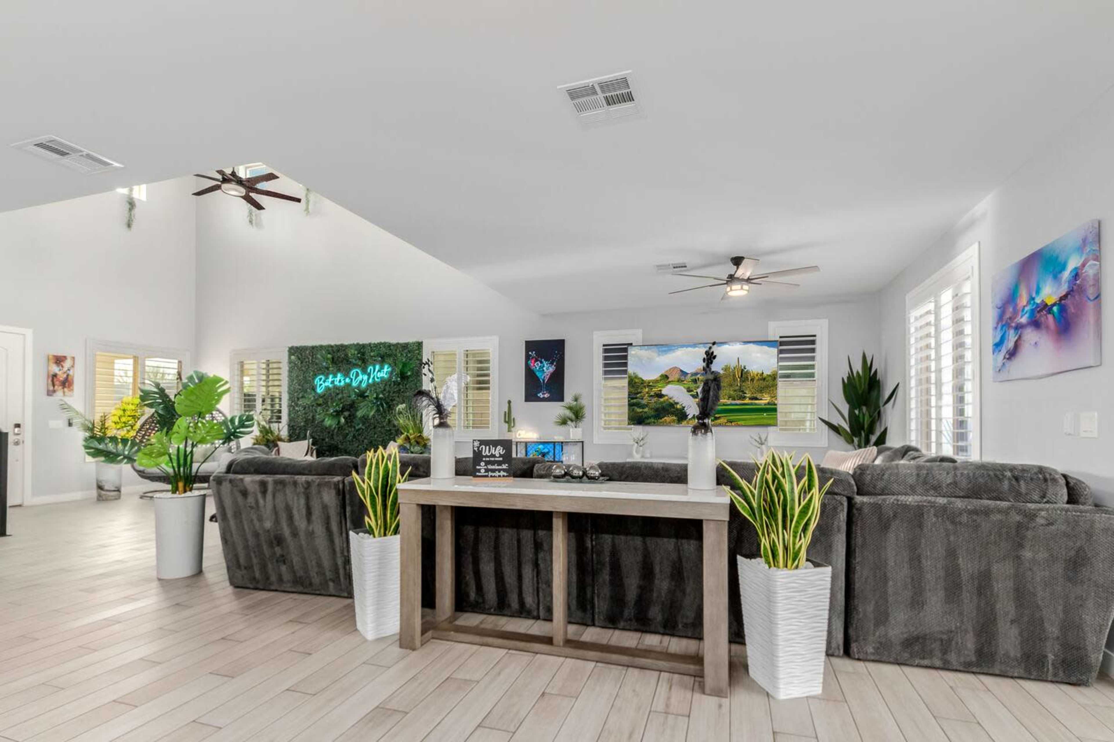 The image shows a modern living room with a large sectional sofa, indoor plants, and a television on a wall, all set in a brightly lit, minimalist space with light-colored flooring.