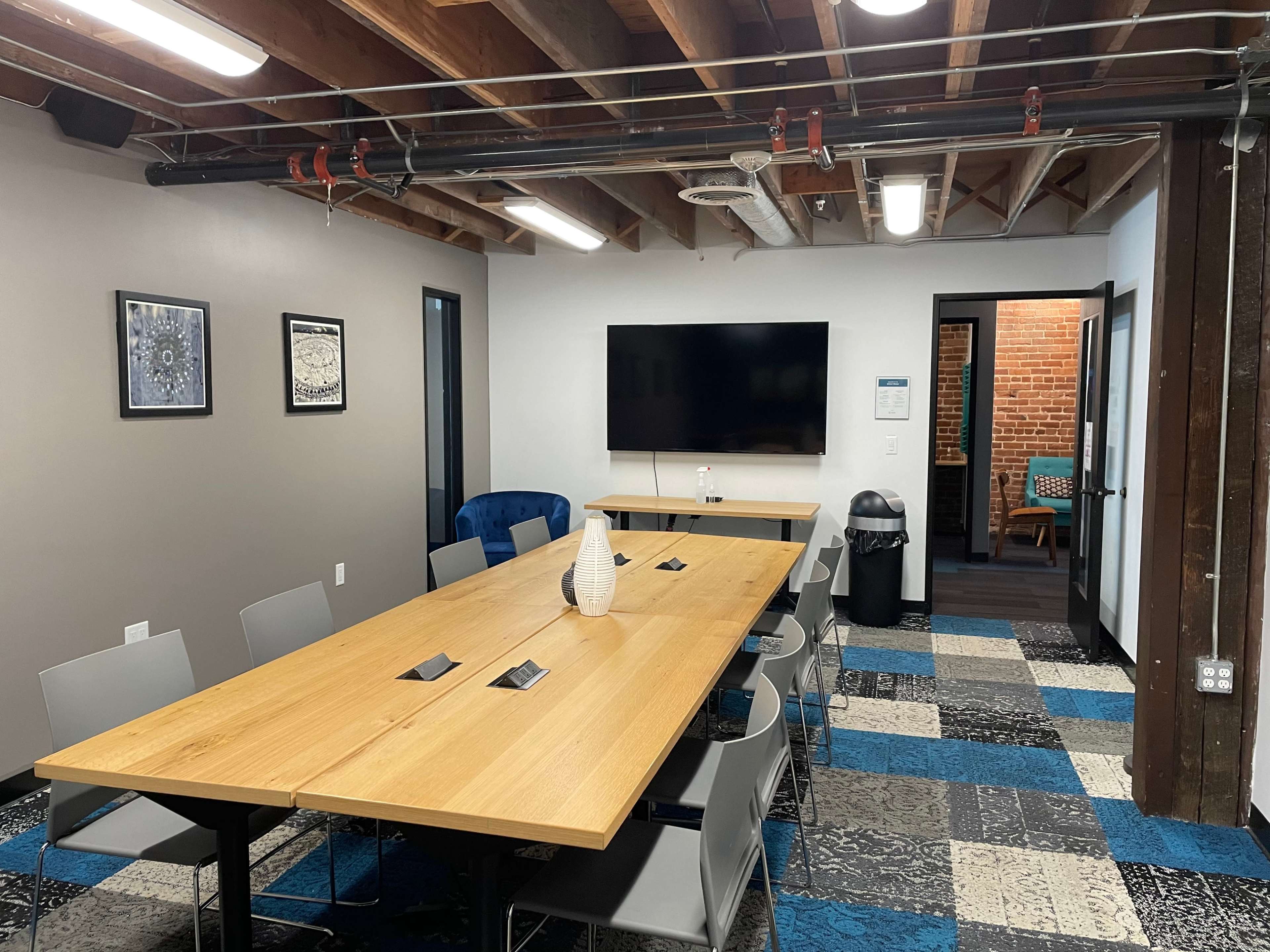 A modern meeting room features a long wooden table surrounded by chairs, a large wall-mounted screen, and exposed beams in the ceiling.