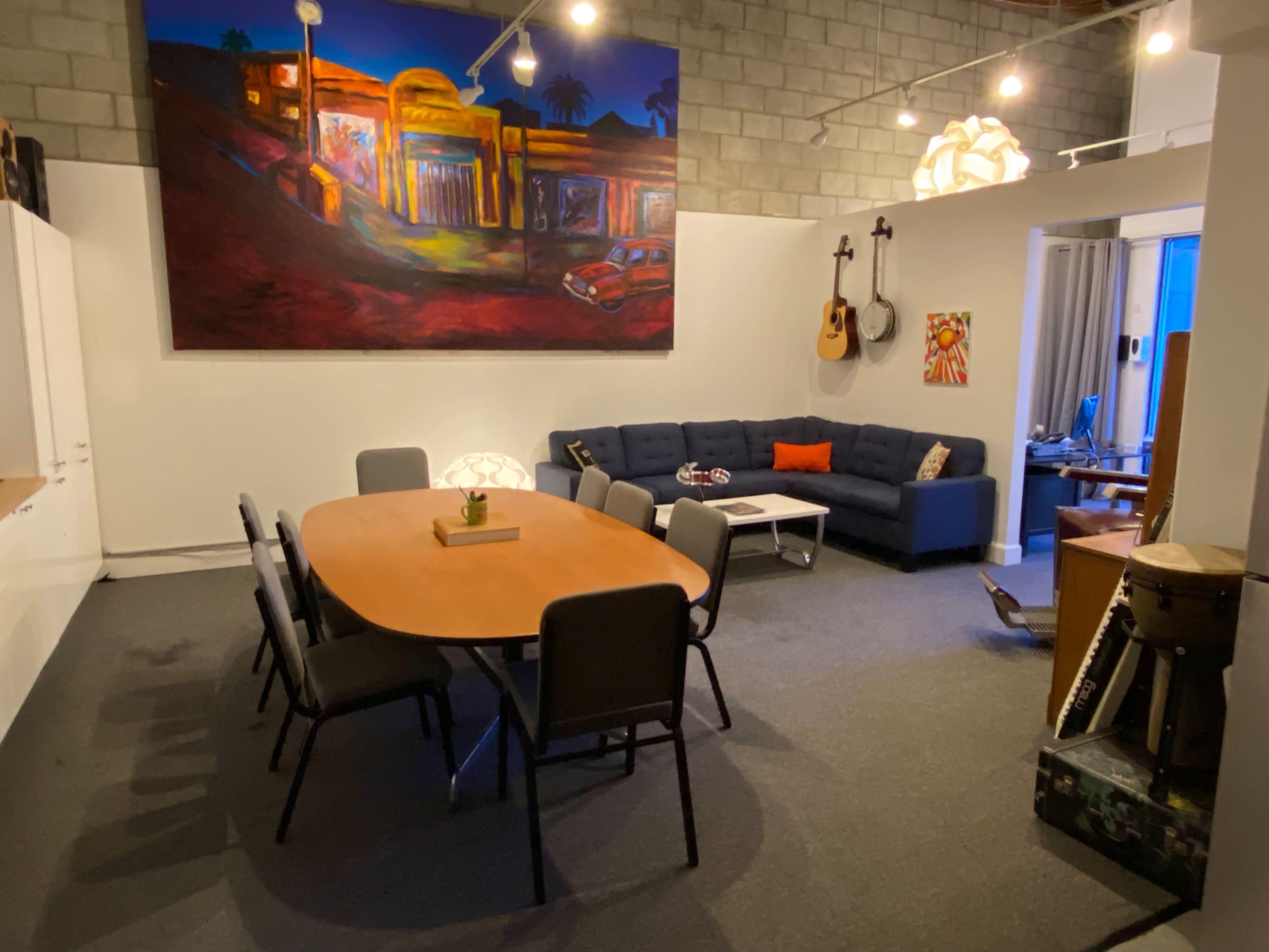 A modern meeting space featuring a large oval table surrounded by chairs, a blue sectional sofa, an artwork on the wall, and various musical instruments.