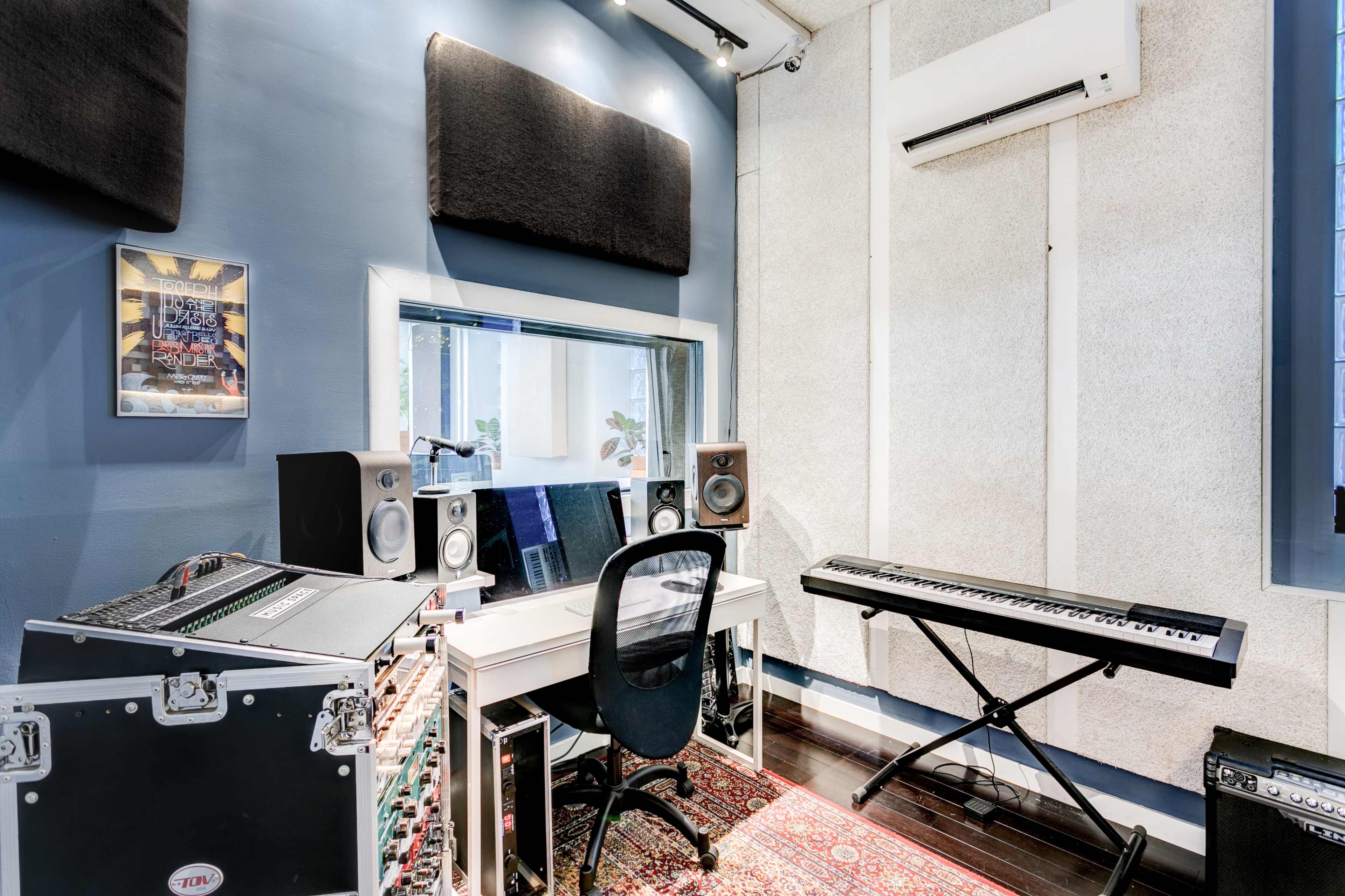 A compact music studio featuring a desk with a computer, audio speakers, a keyboard, and soundproofing panels on the walls.