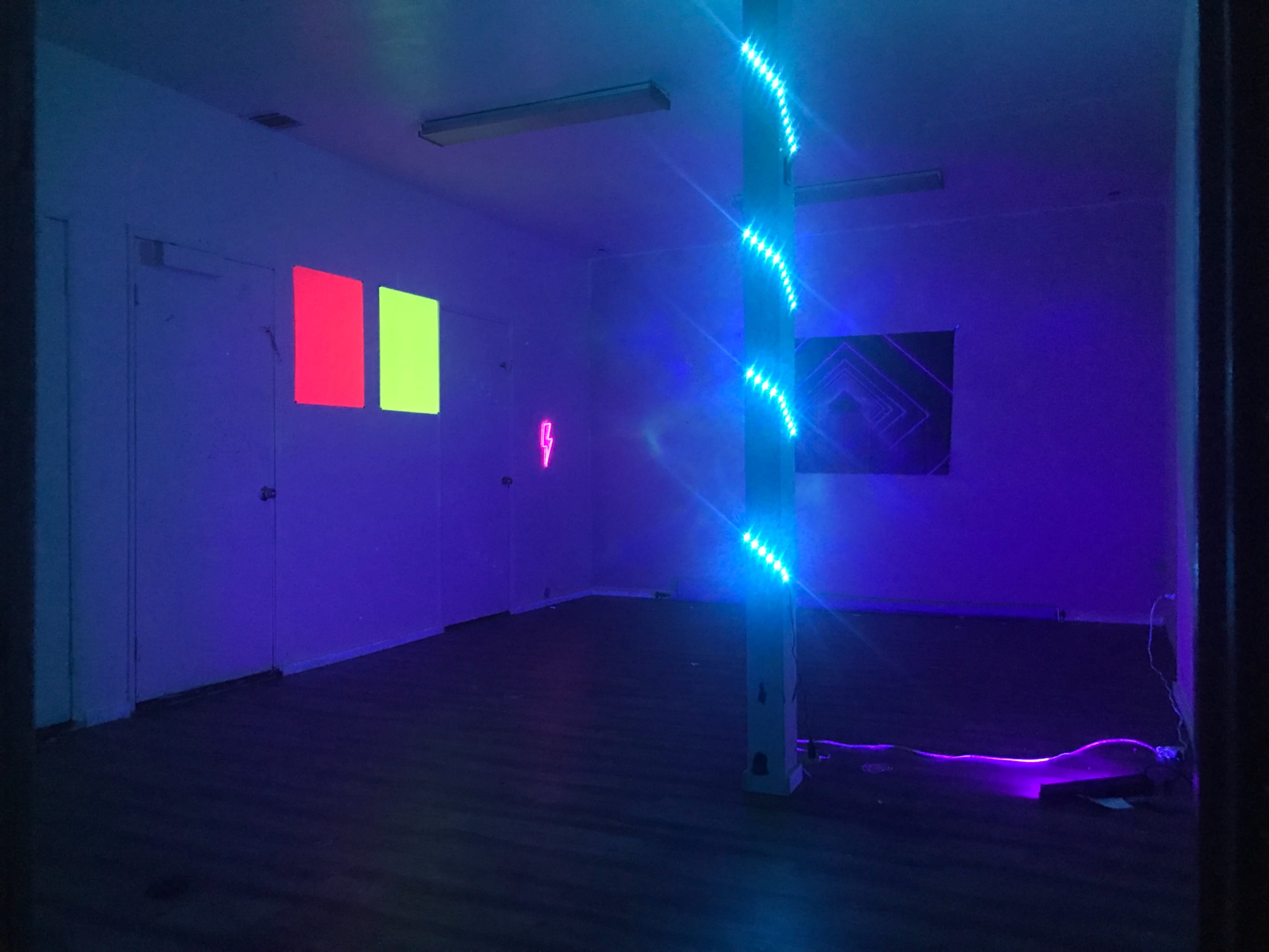 An empty room with a light blue pillar entwined with LED lights, two colored squares projected on the wall, and a neon light design in the background.